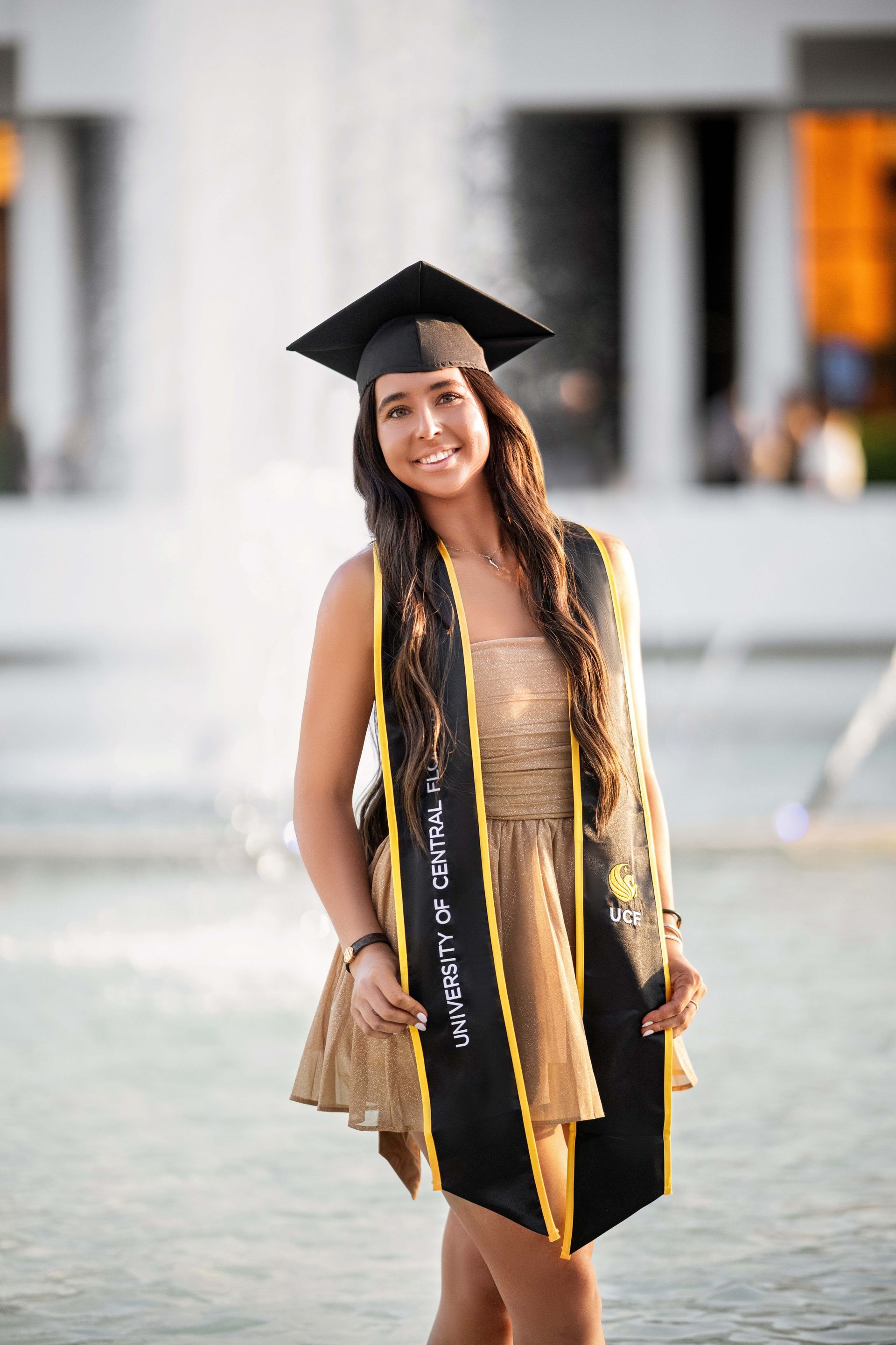 Woman in graduation attire smiles near a fountain. She wears a black cap, gold dress, and sash.