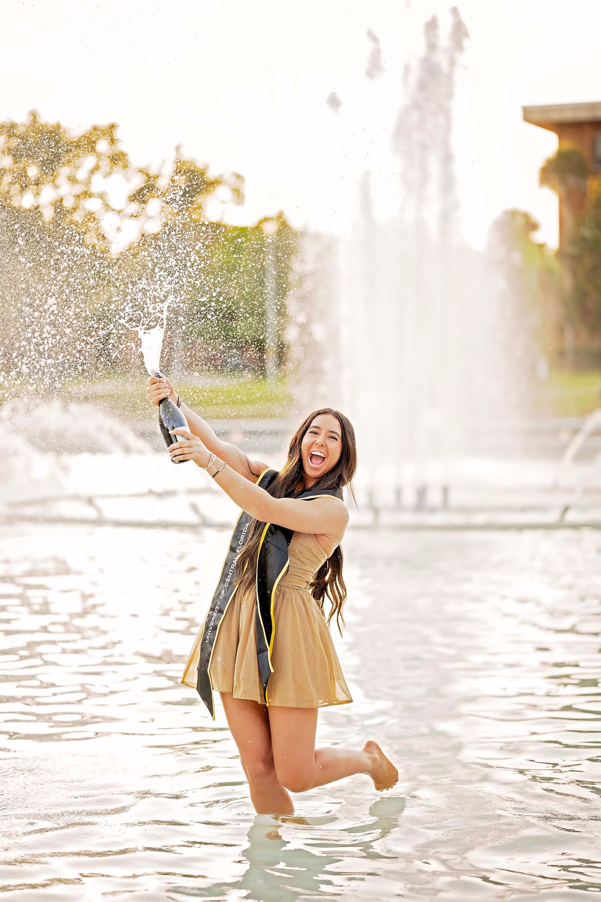 Woman celebrating graduation in a fountain, popping champagne with water spraying up.