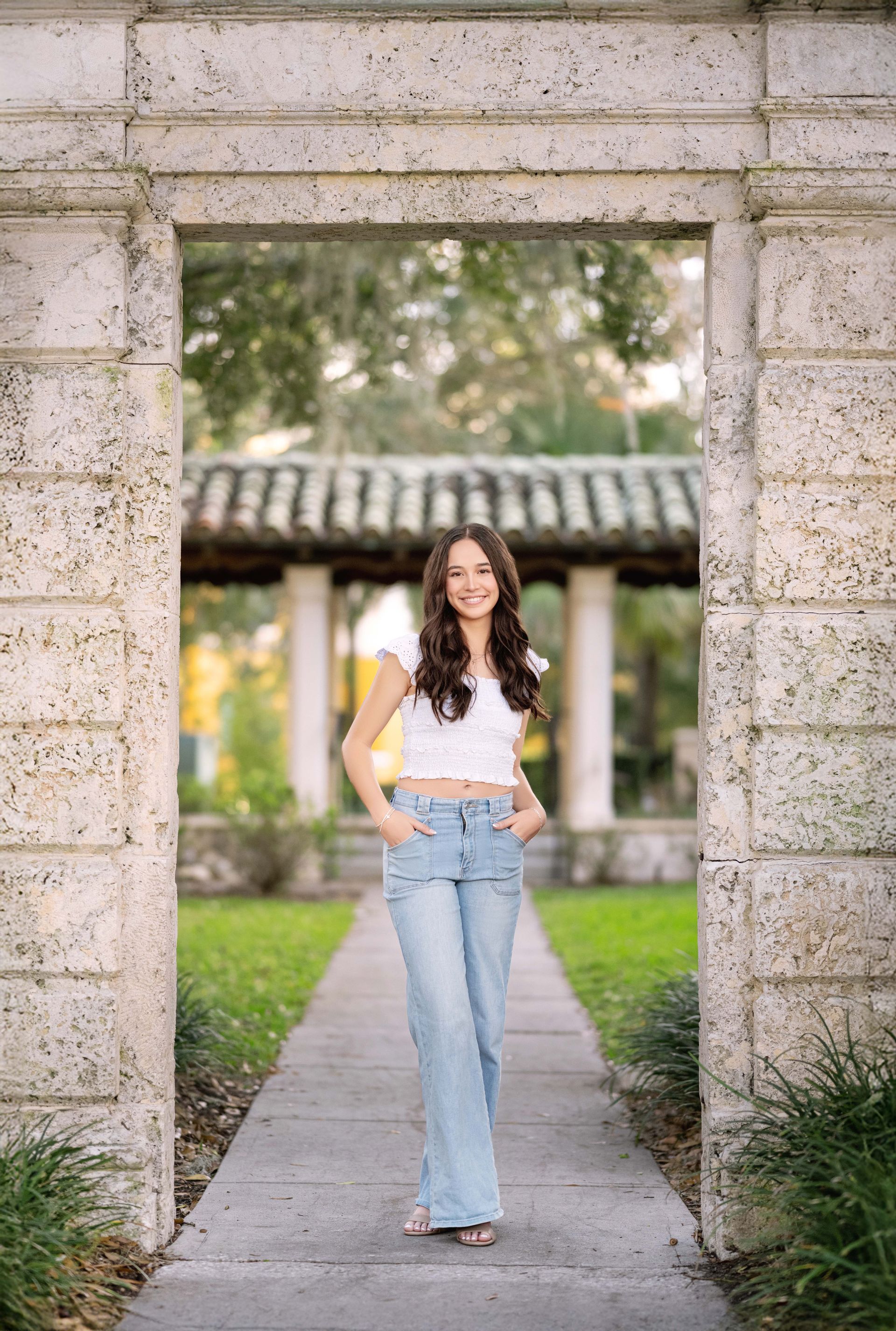 Young woman in jeans and crop top smiles, standing in stone archway, garden in background.