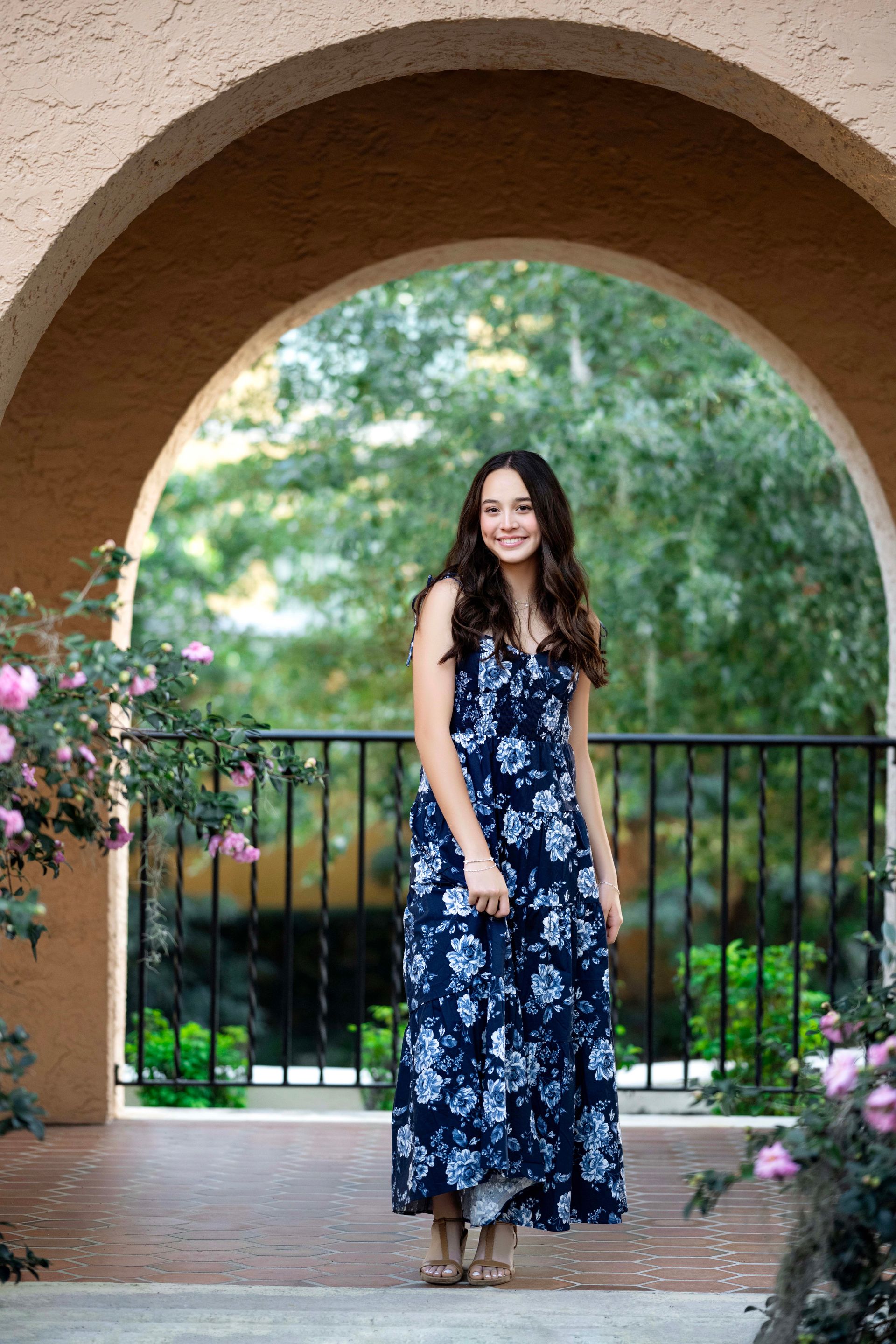 Woman in floral dress, smiling, standing under an archway. Flowers and greenery in the background.