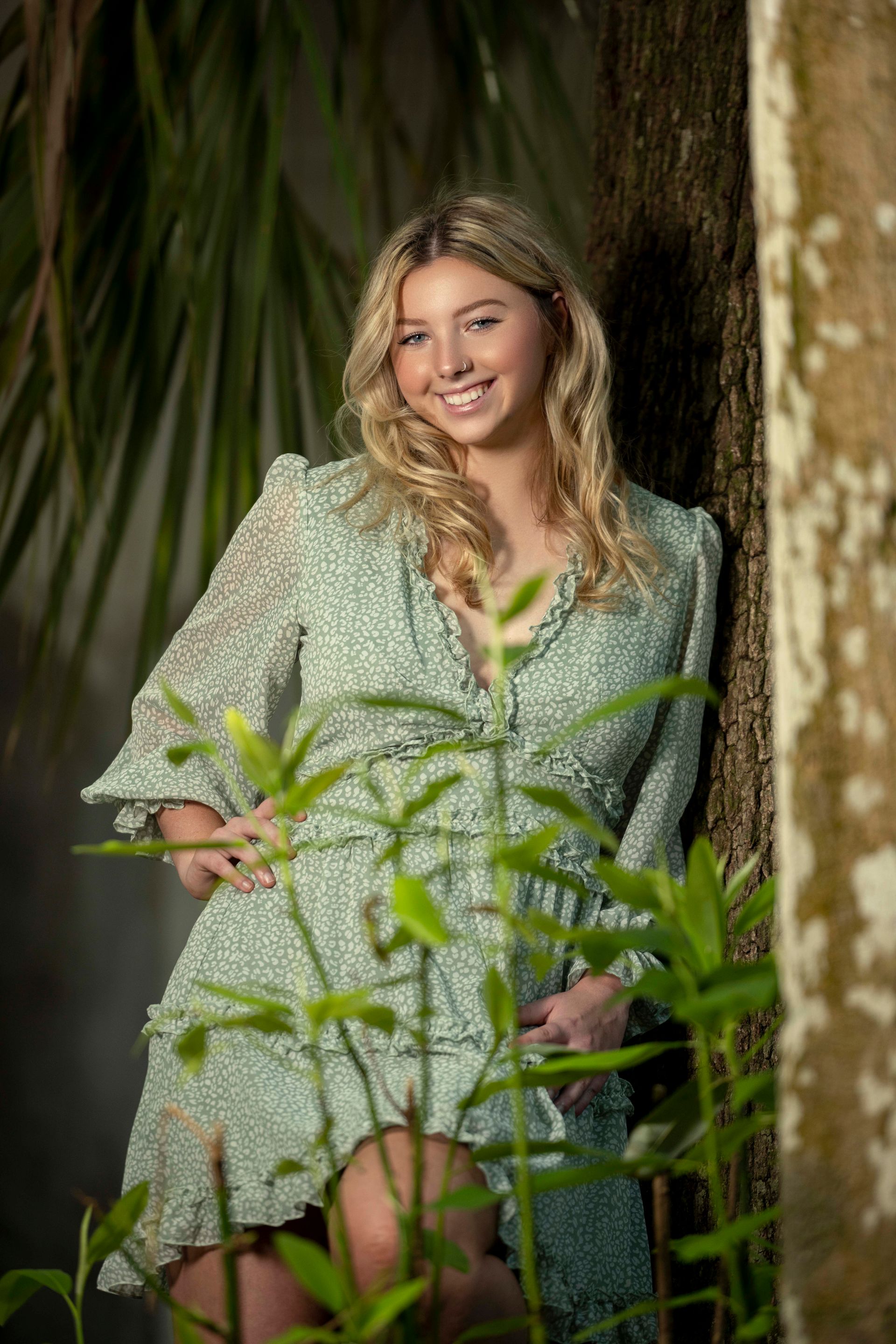 Blonde woman in green dress smiles, leaning against a tree outdoors.