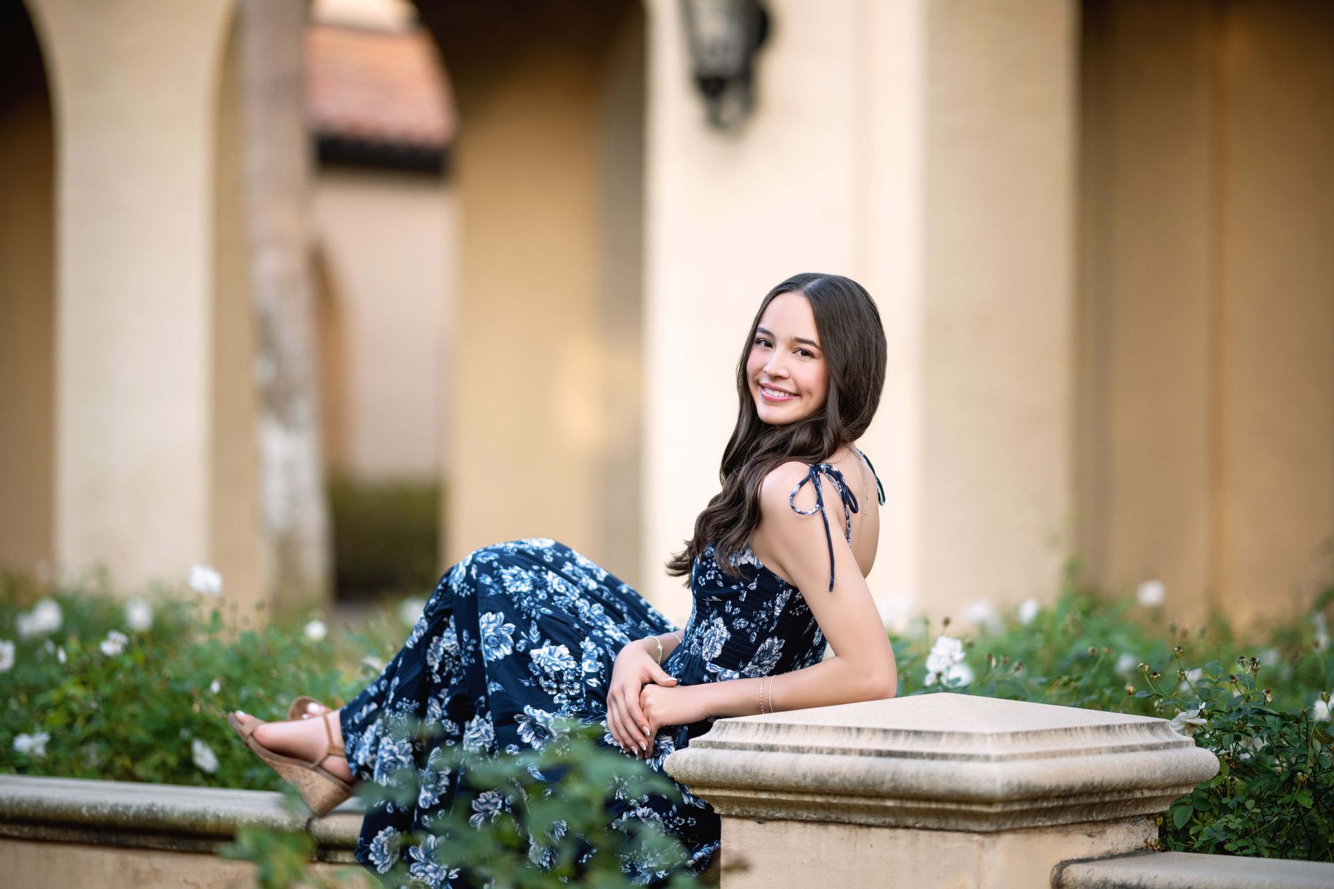 Young woman in blue floral dress smiles, sits on a stone ledge with flowers, outdoors.