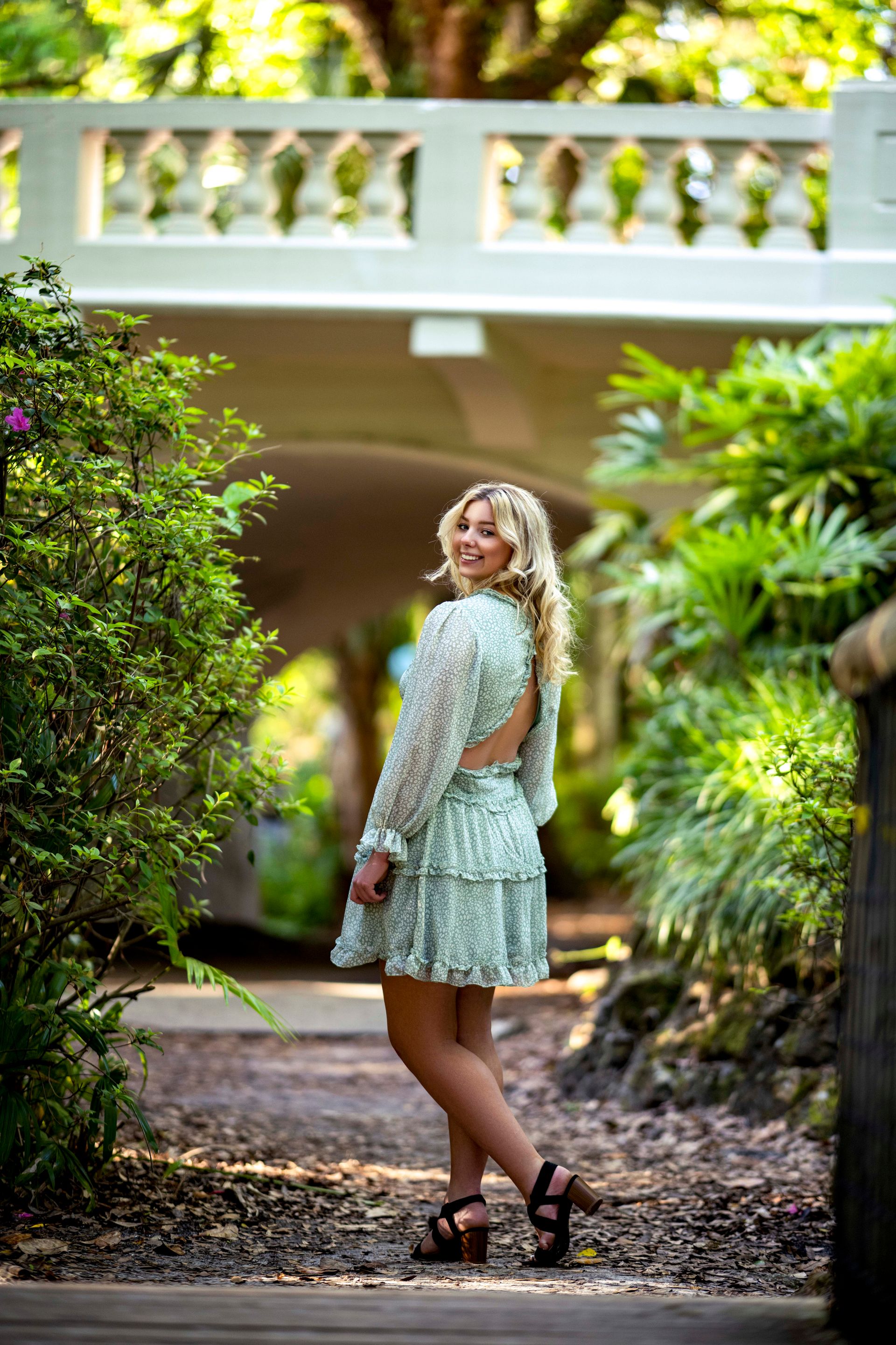 Blonde woman in a green dress smiles, turned towards the camera, under a white bridge in a garden.