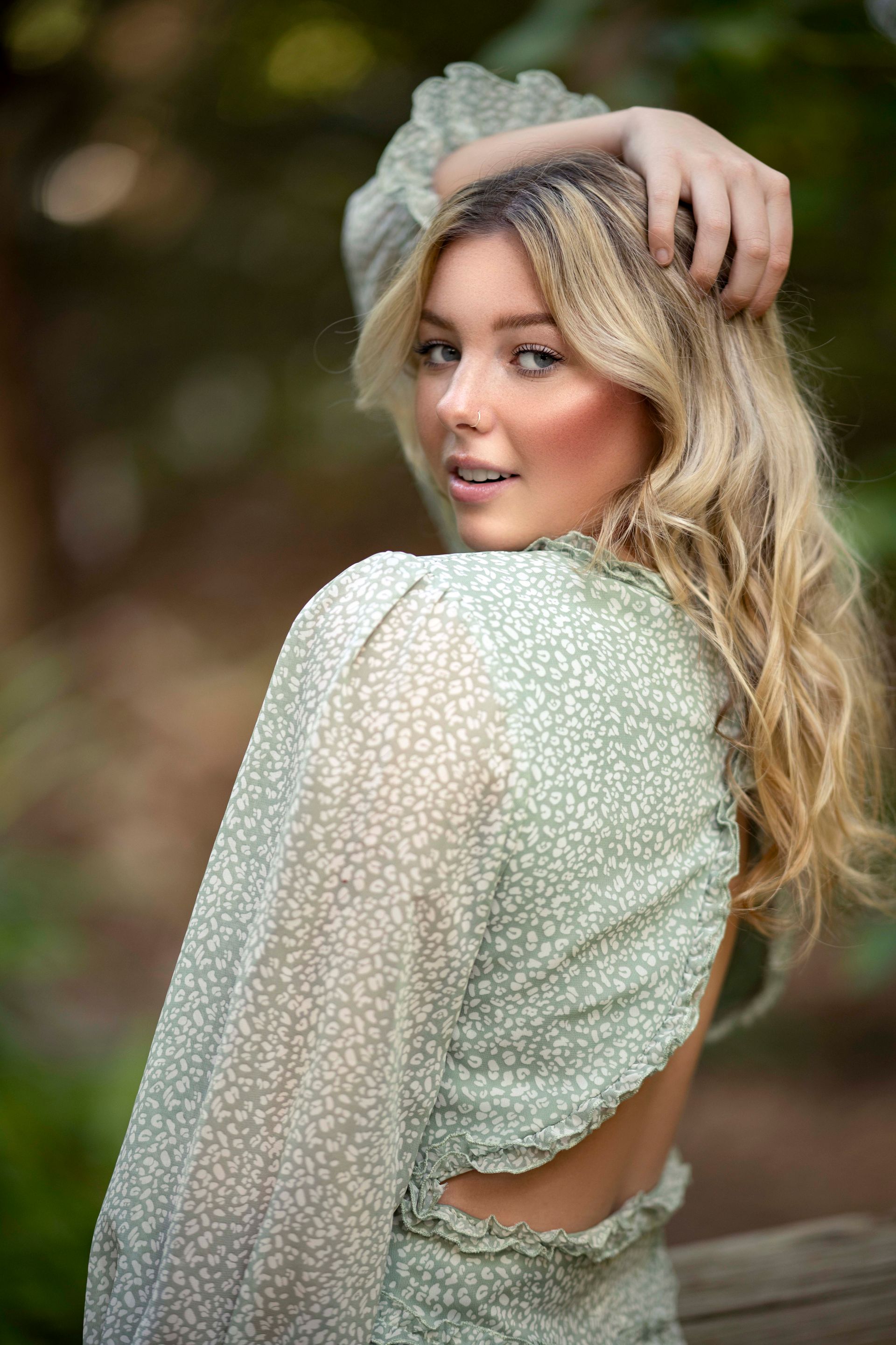 Blonde woman in a green dress, posing outdoors, smiling, touching her hair.
