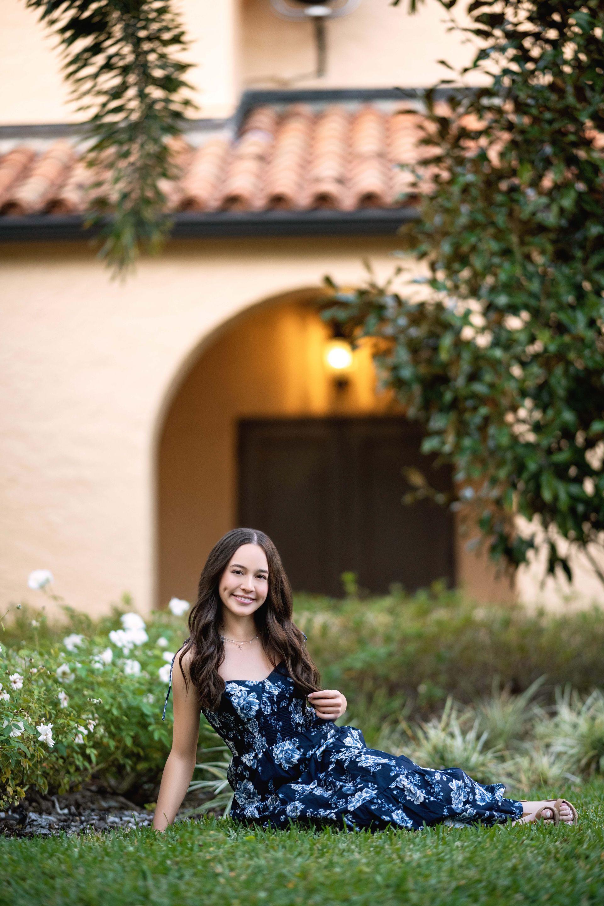 Young woman in floral dress sitting on grass, smiling, in front of a building with an arched doorway.