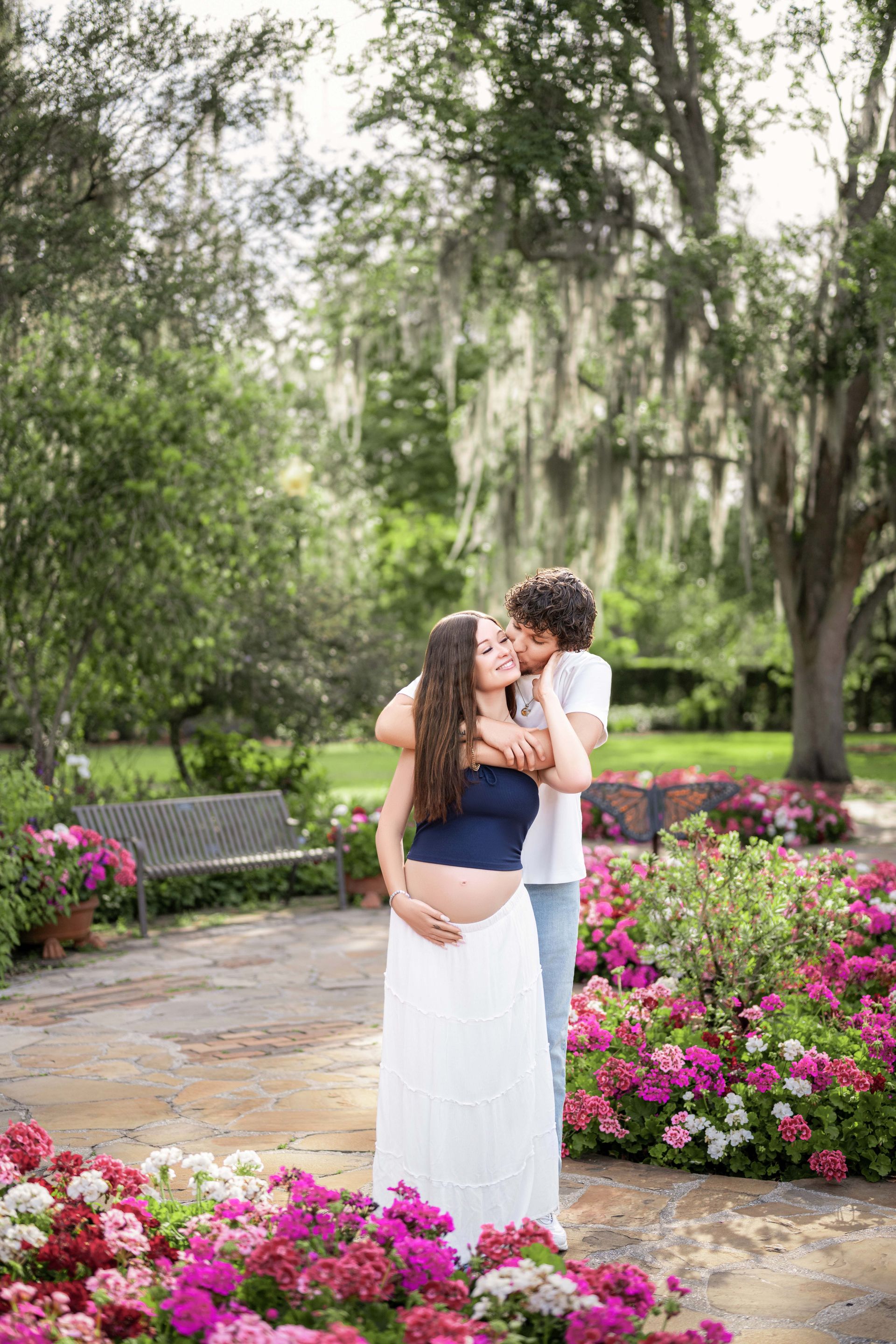 Pregnant woman and partner embrace among flowers in a park.