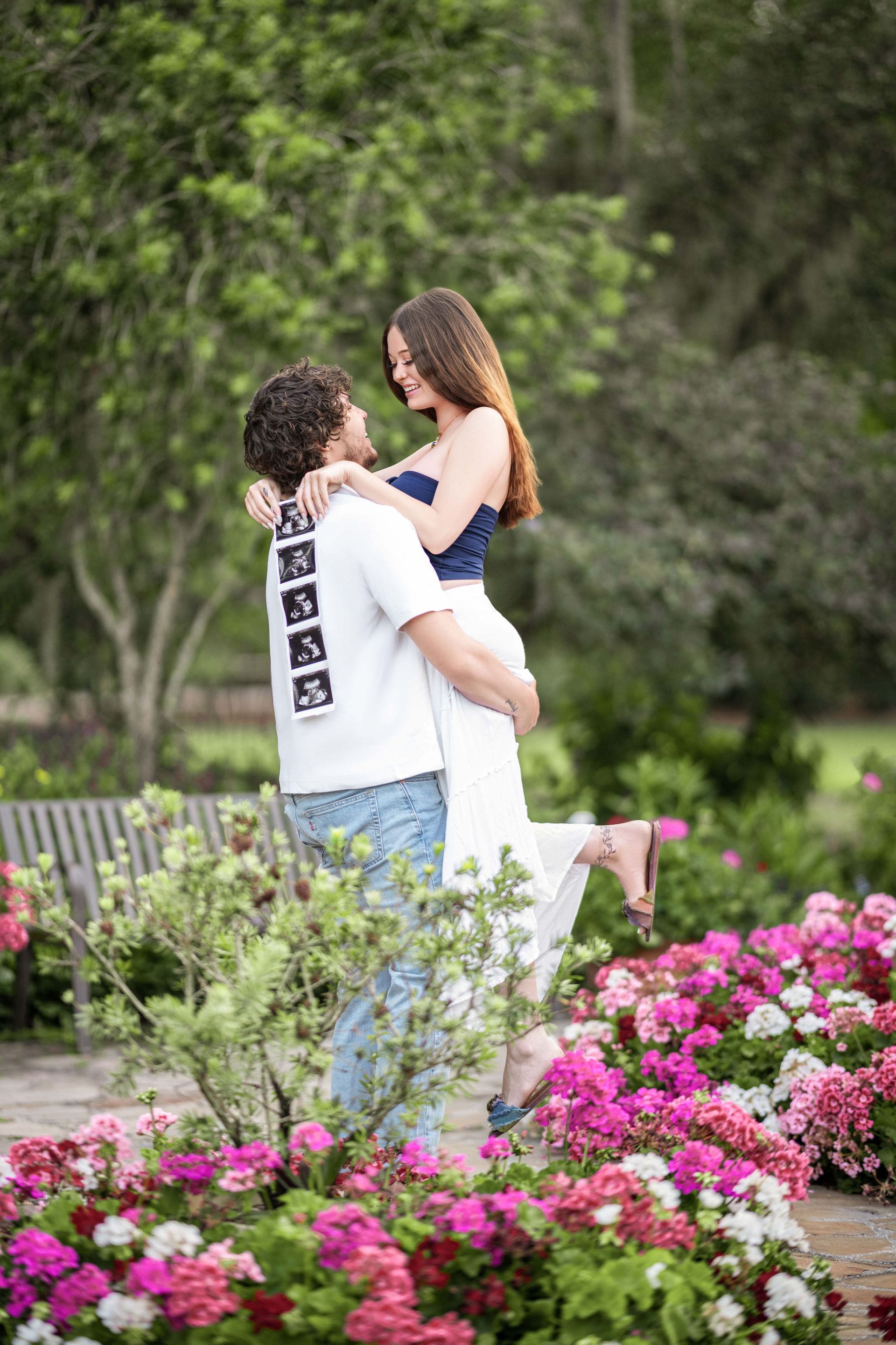Man lifting woman, surrounded by flowers and greenery. They face each other smiling in a garden setting.