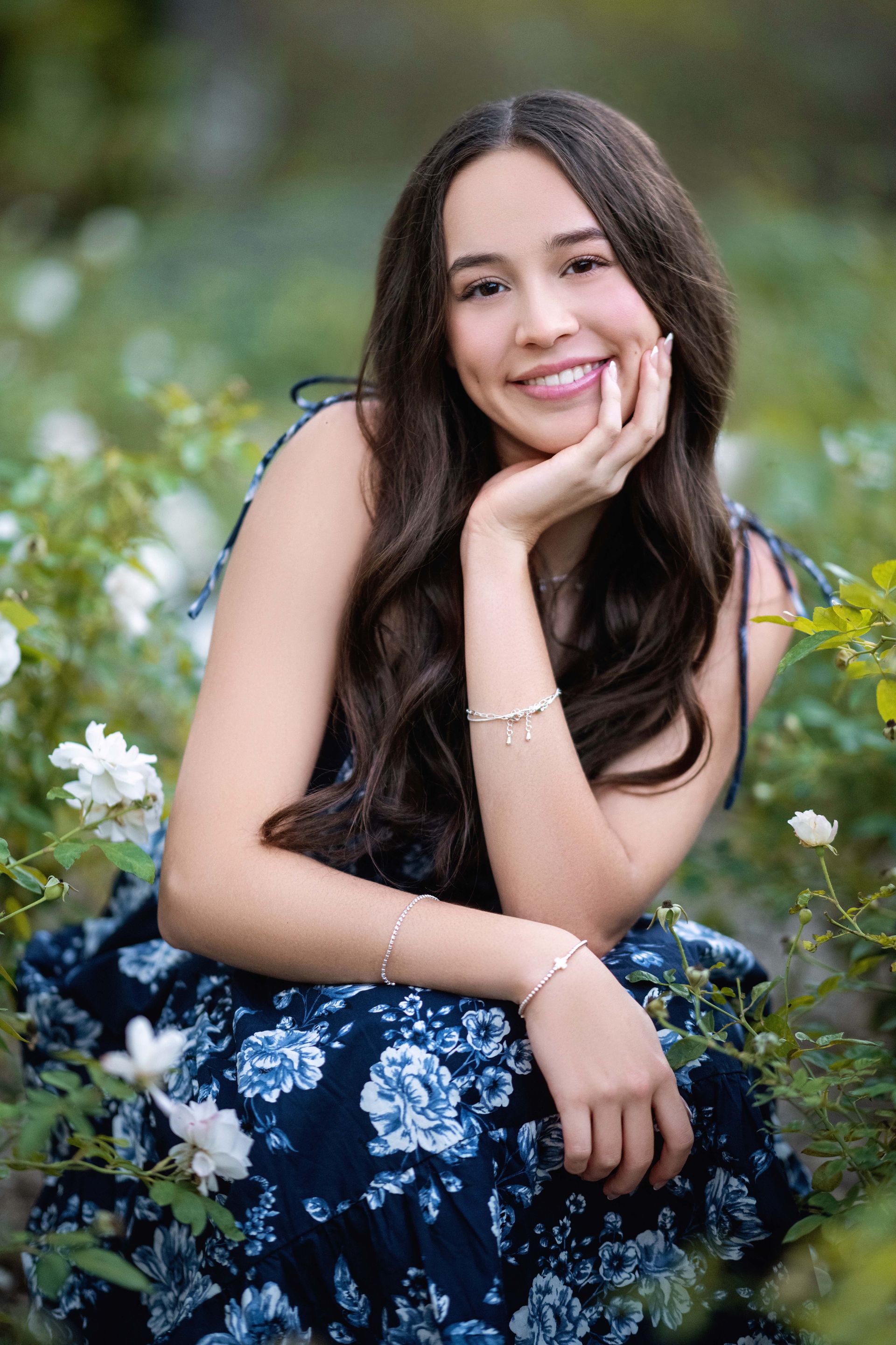 Young woman smiling, hand on chin, surrounded by white flowers, wearing blue floral dress.