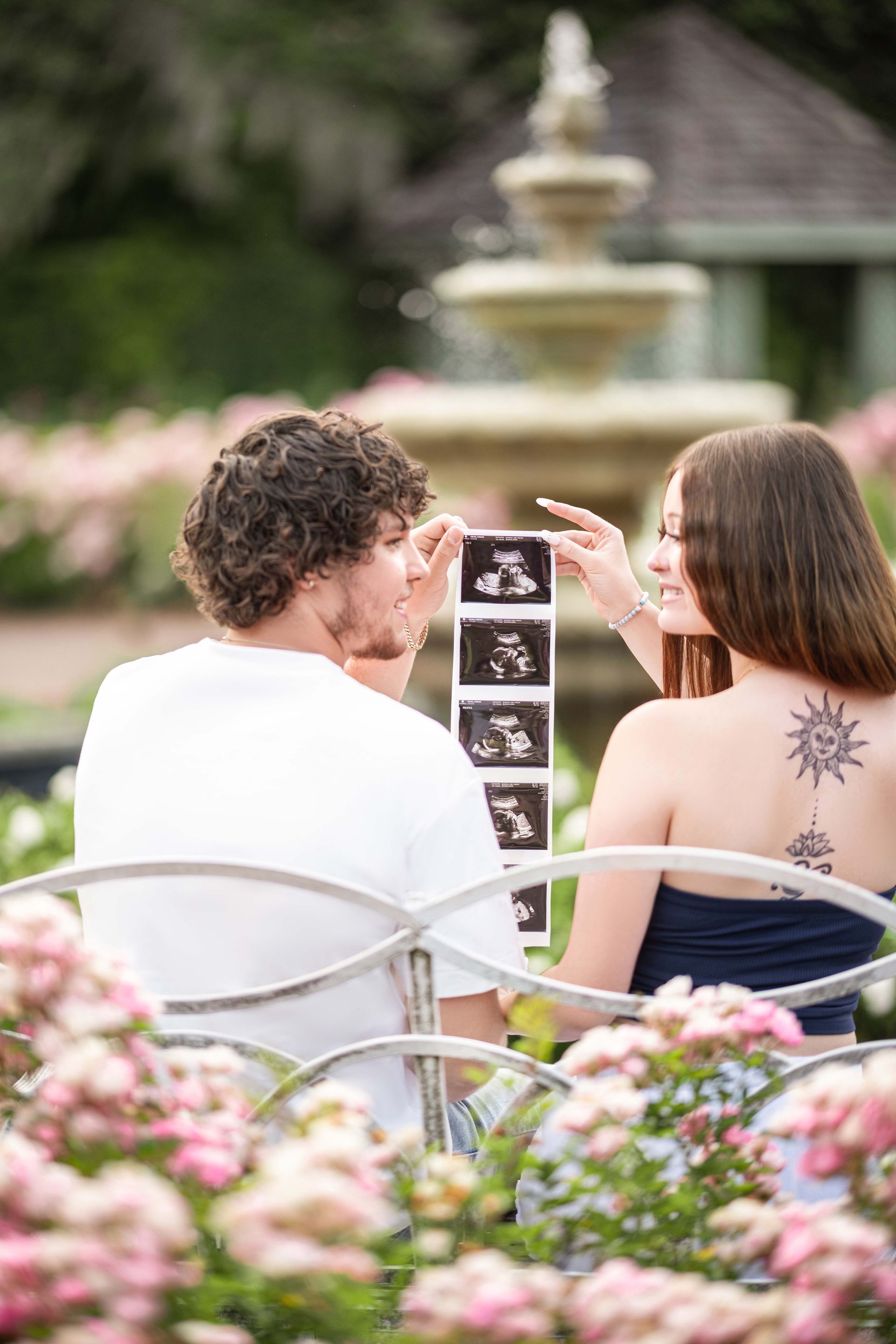 Couple in garden holding ultrasound images; fountain and flowers in background.