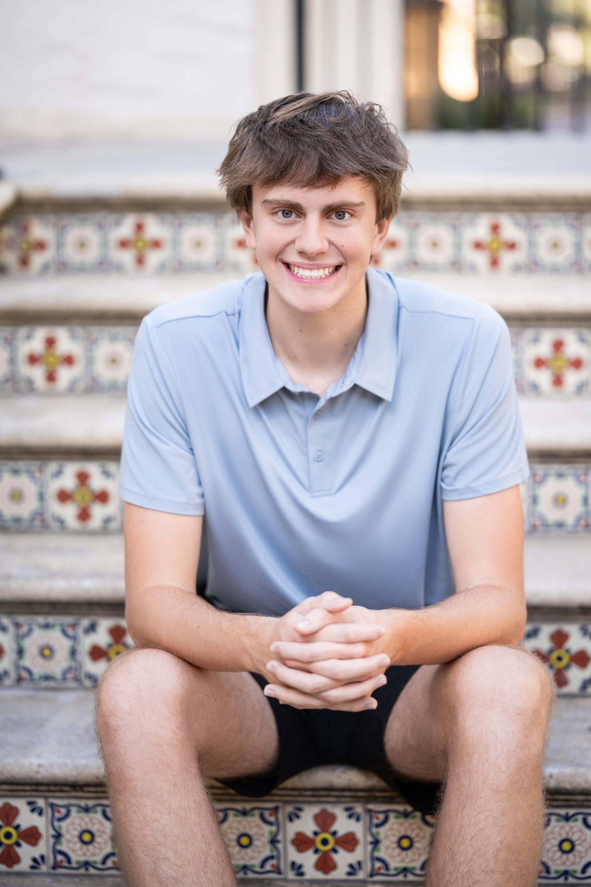 Young man with curly brown hair smiling, sitting on steps, wearing a blue polo shirt and black shorts.