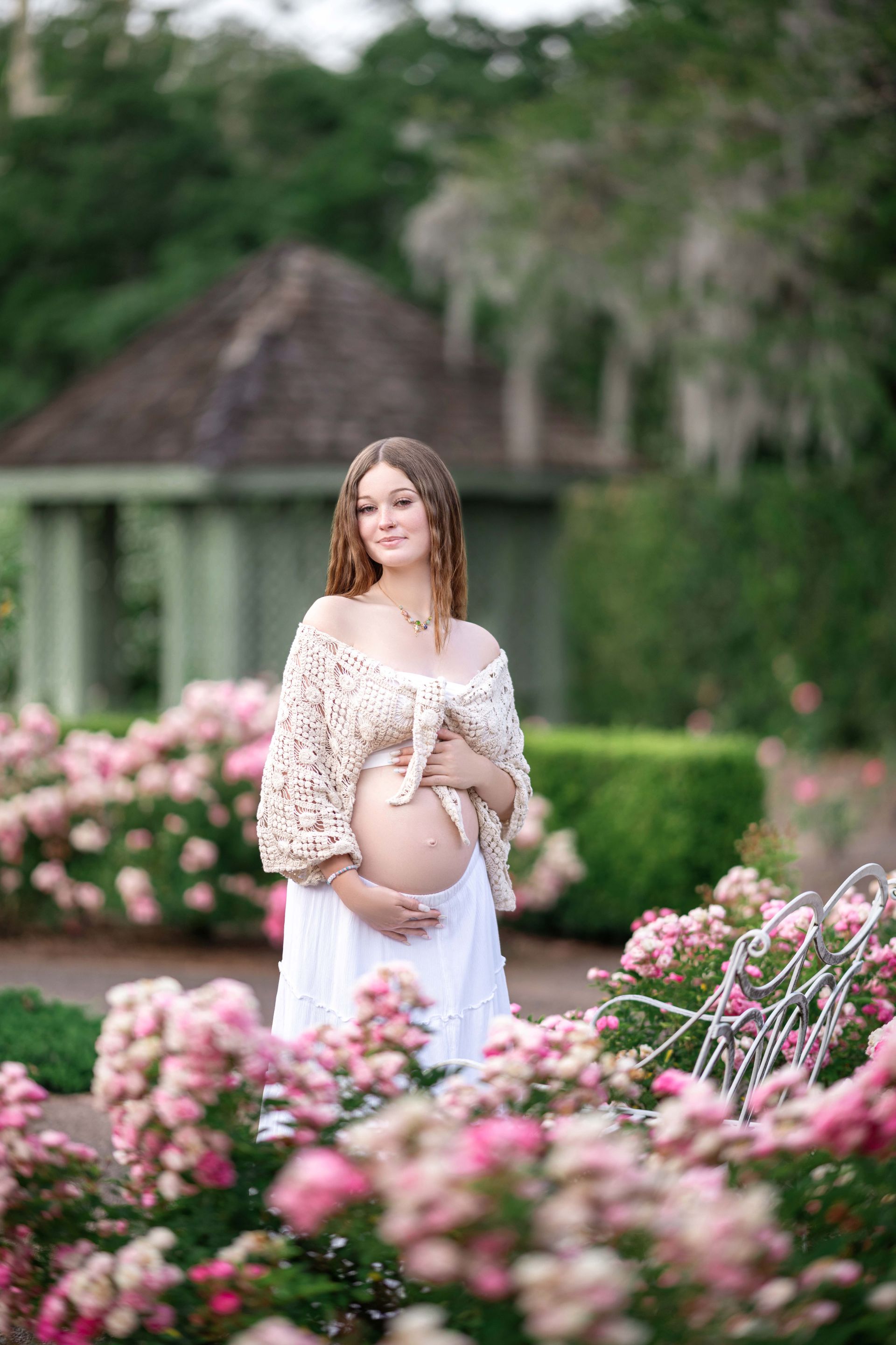 Pregnant woman in a garden, wearing a beige shawl, embraces her belly. Surrounded by pink roses.