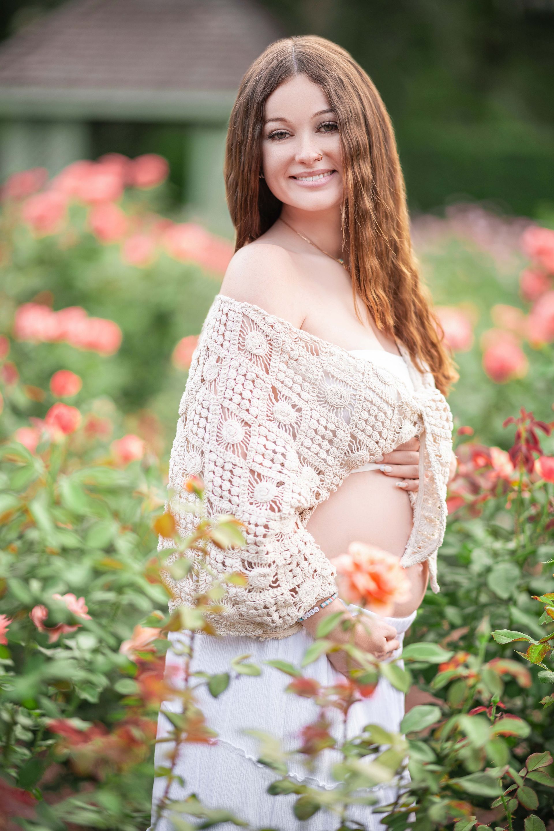 Pregnant woman smiling in a rose garden, wearing a shawl, holding her belly.
