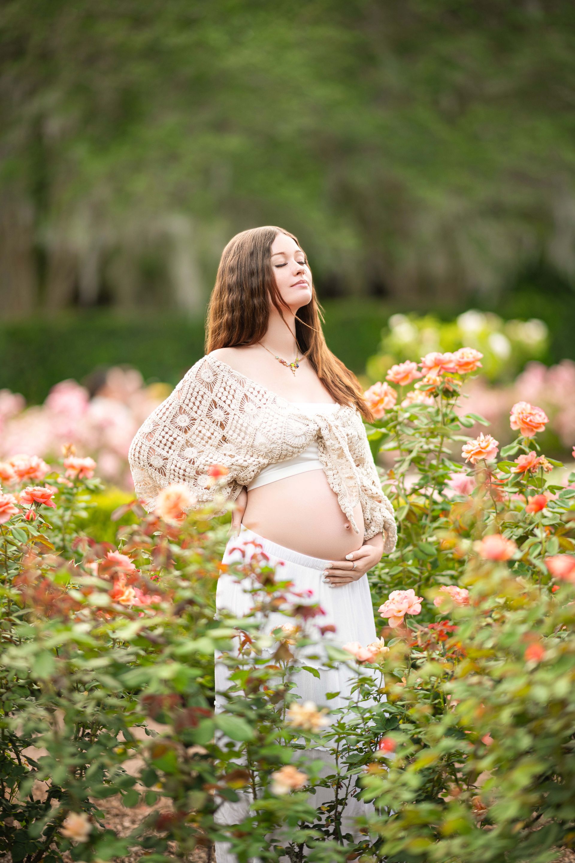 Pregnant woman in white outfit and floral wrap, outdoors in a rose garden, holding her belly.