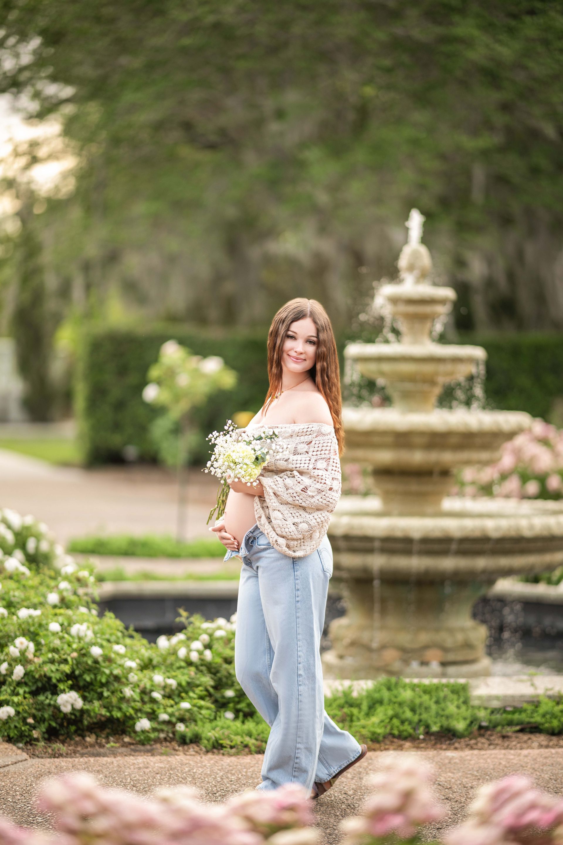 Pregnant woman in jeans and off-the-shoulder top, holding flowers, smiles in a garden with a fountain.