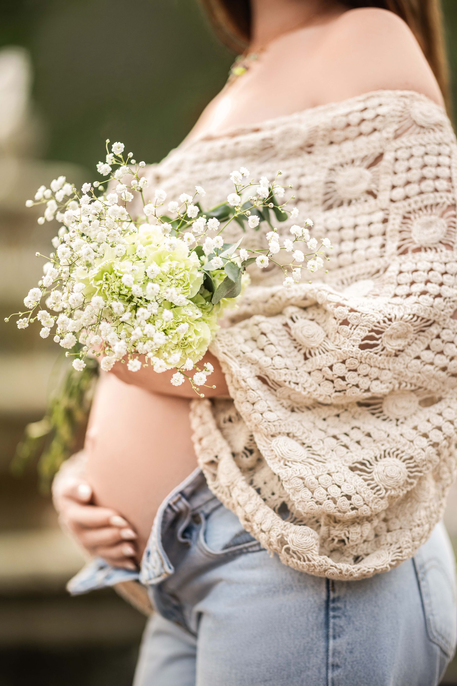 Pregnant woman holding flowers, wearing a crocheted shawl and denim overalls.