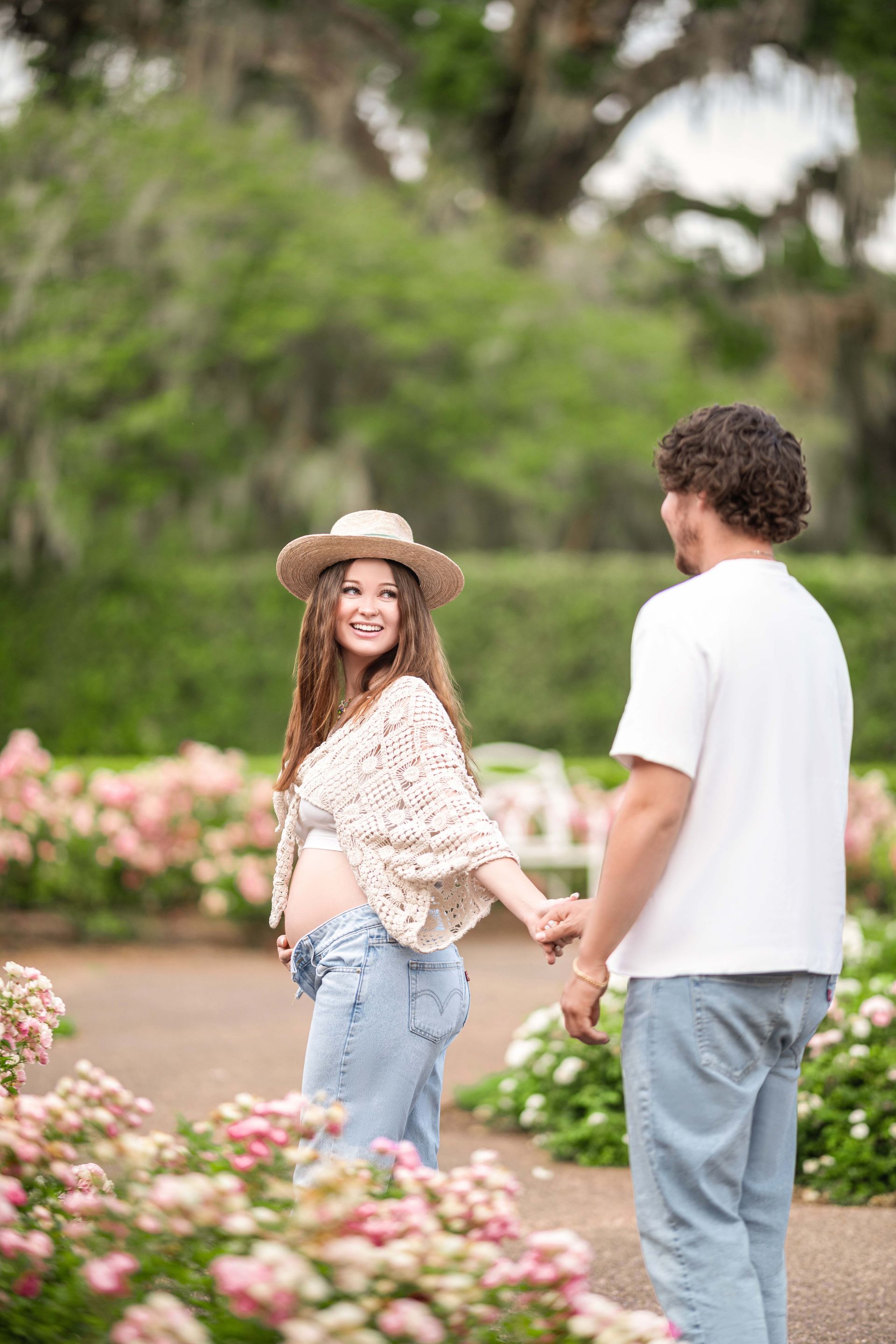 Pregnant woman and man holding hands, smiling in a garden with pink flowers.