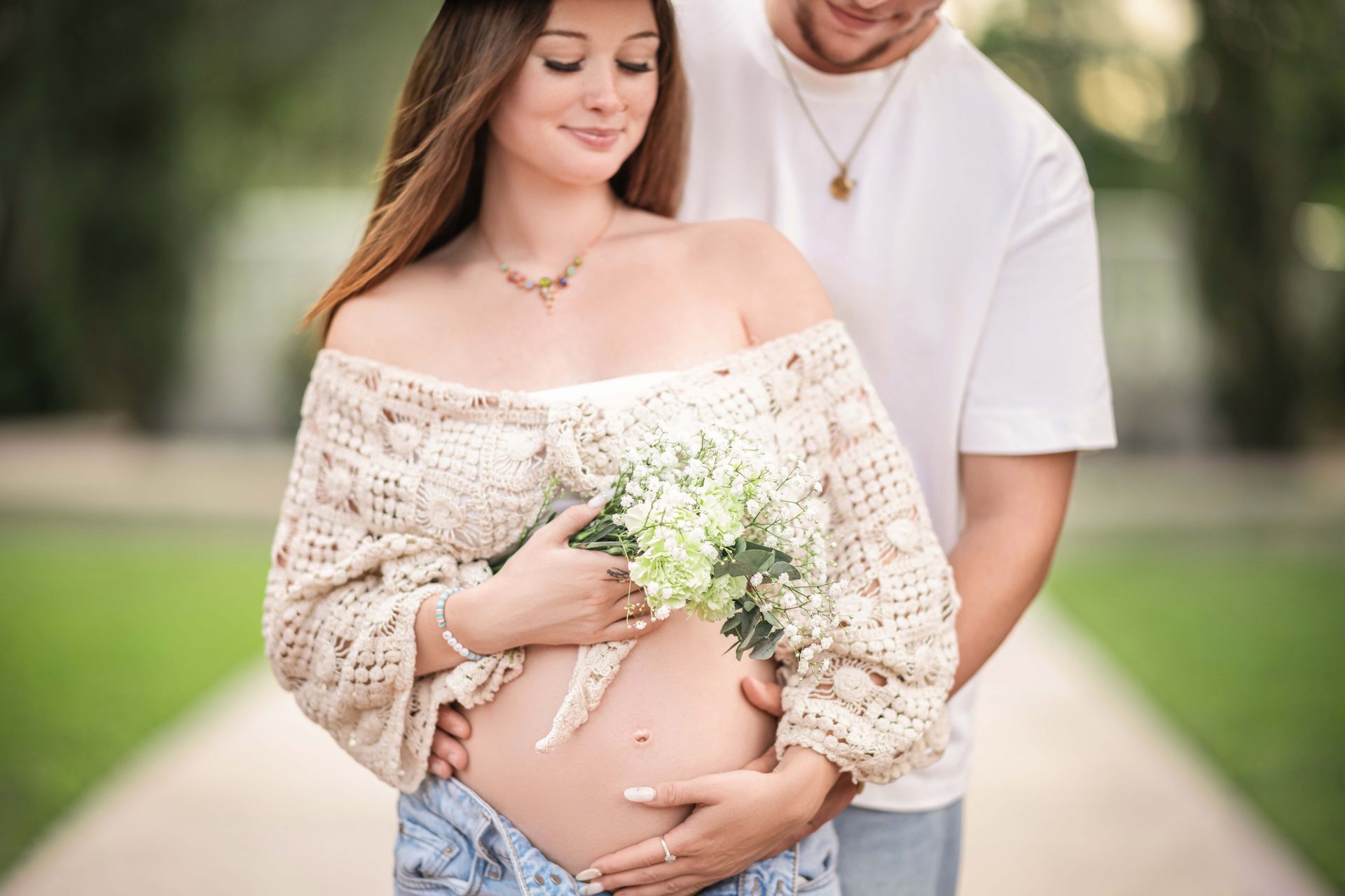 Pregnant woman and partner in outdoor setting. Woman cradles belly, holding flowers.