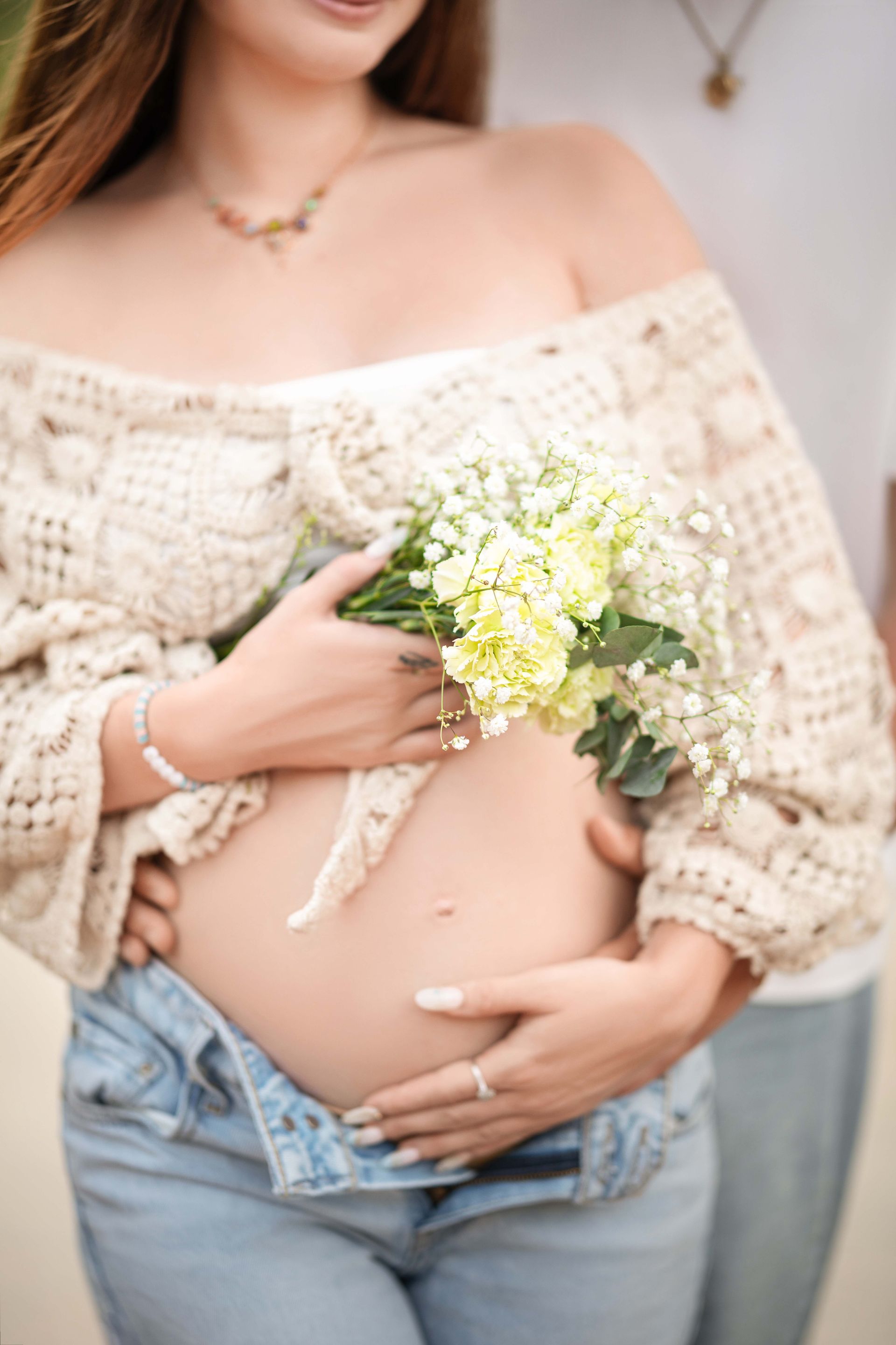 Pregnant woman holds bouquet, embraces belly; another person's arms around her.