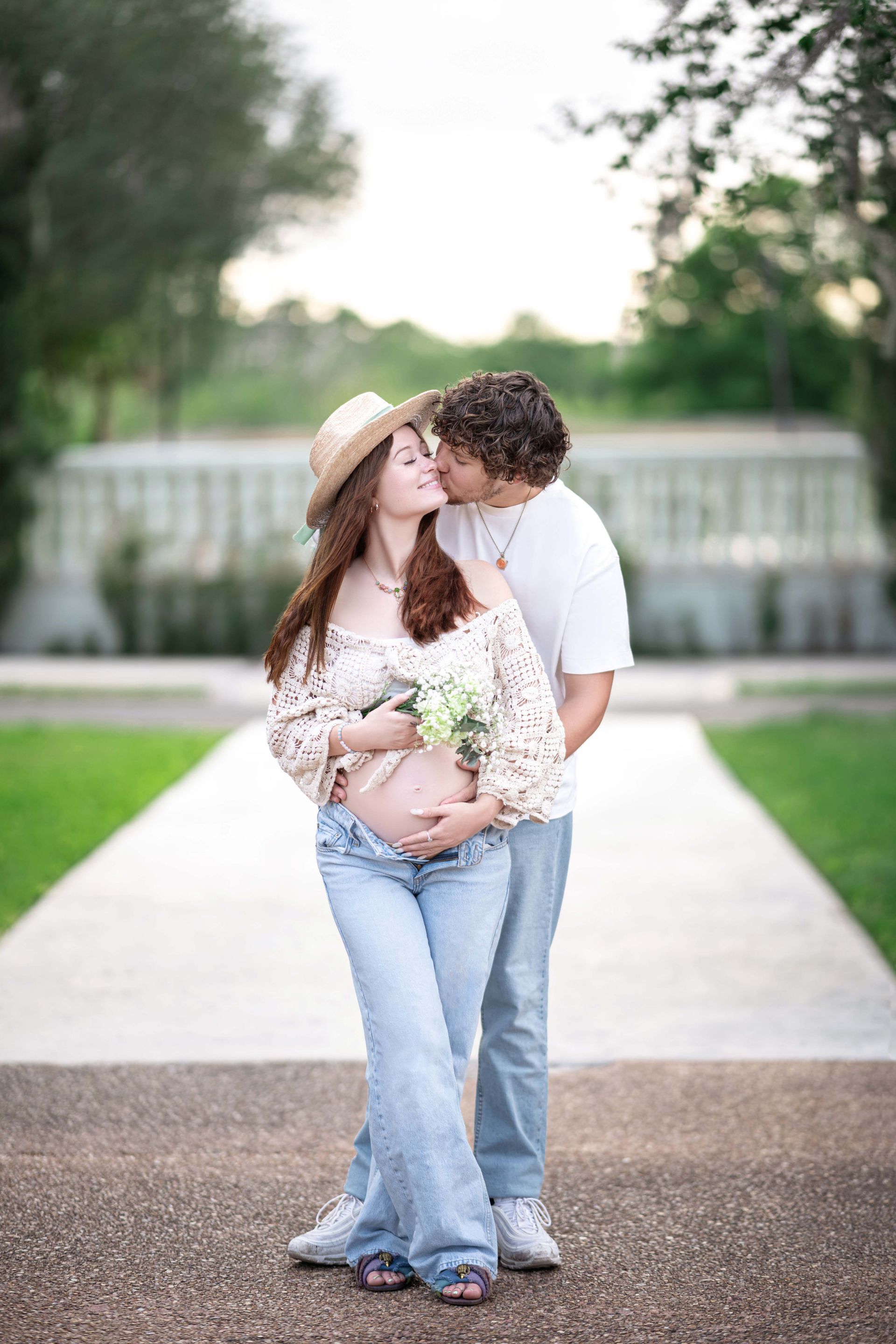 Pregnant couple kissing outdoors, holding baby bump. Woman wears hat, floral top, and jeans. Man wears white shirt and jeans.
