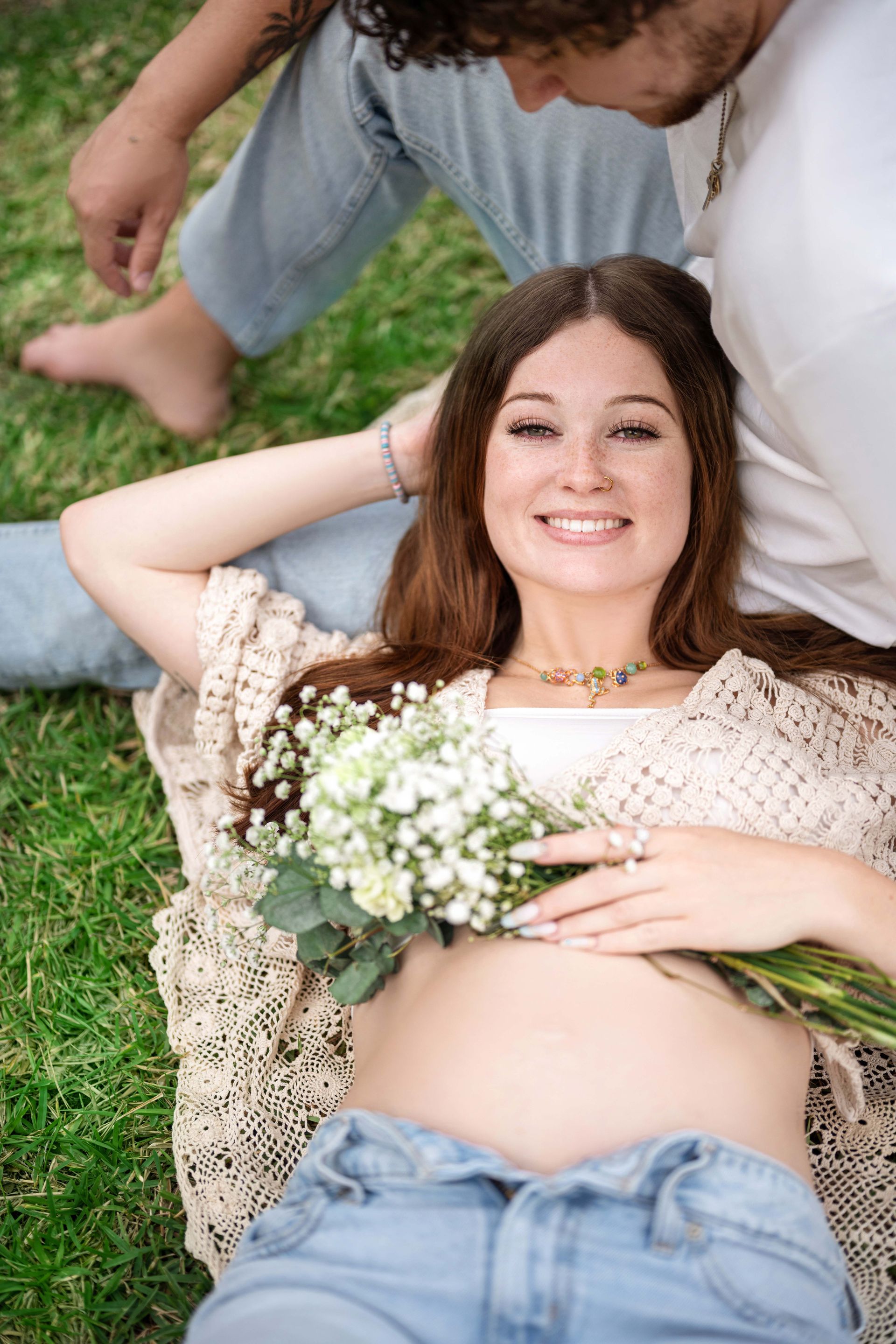 Pregnant woman smiling, lying on grass, holding flowers. Man's legs and arm visible. Sunny, outdoor setting.