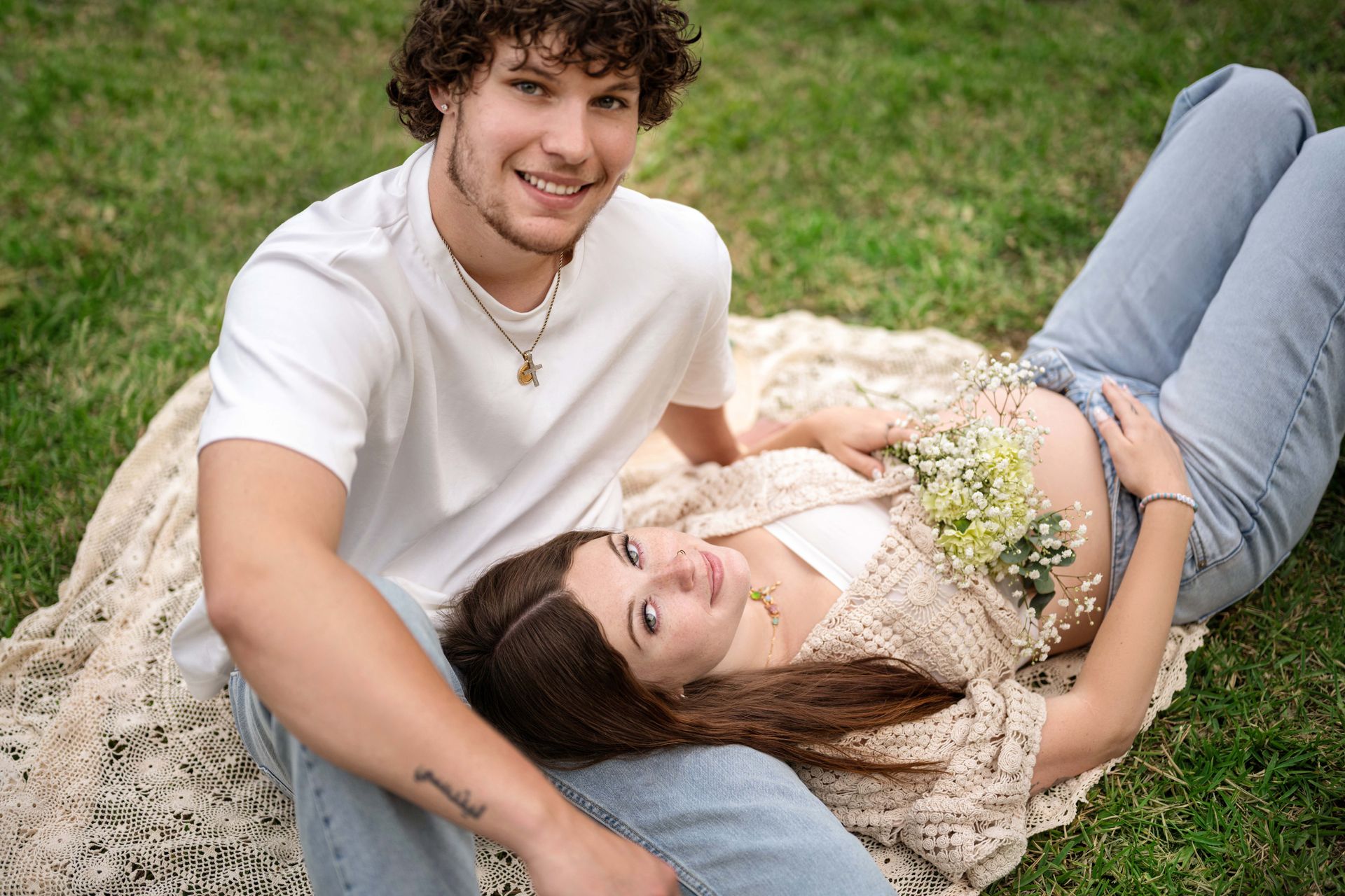 Pregnant woman laying on a blanket in grass, with partner. Both are smiling.
