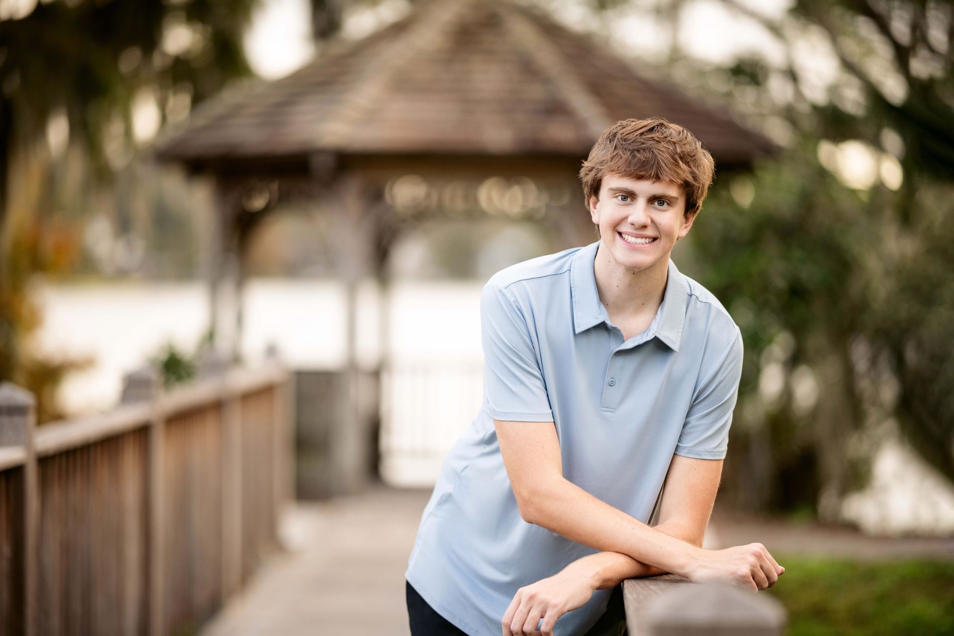 Young man in blue shirt leaning on a wooden railing, smiling near a gazebo and lake.