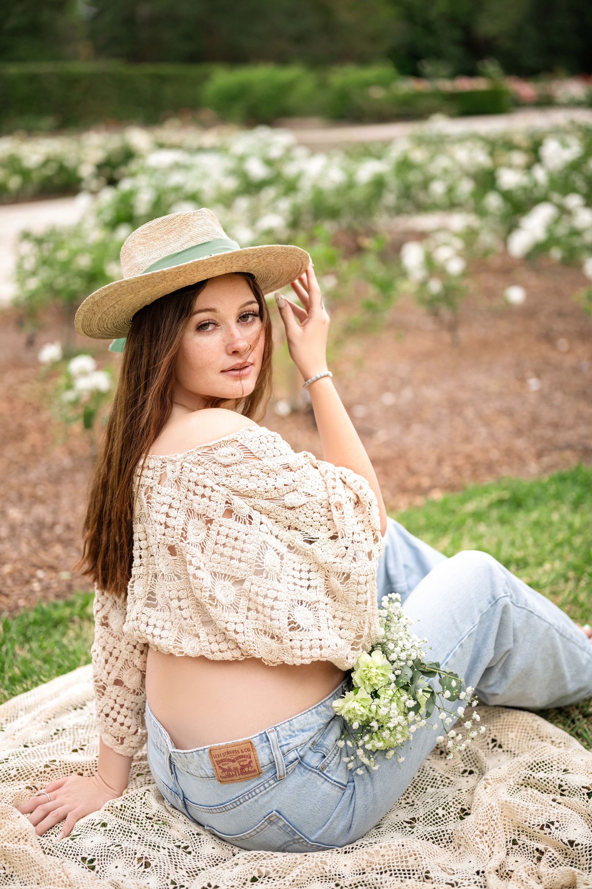 Woman in straw hat and crocheted top, sitting on a blanket, holding flowers, outdoor setting.
