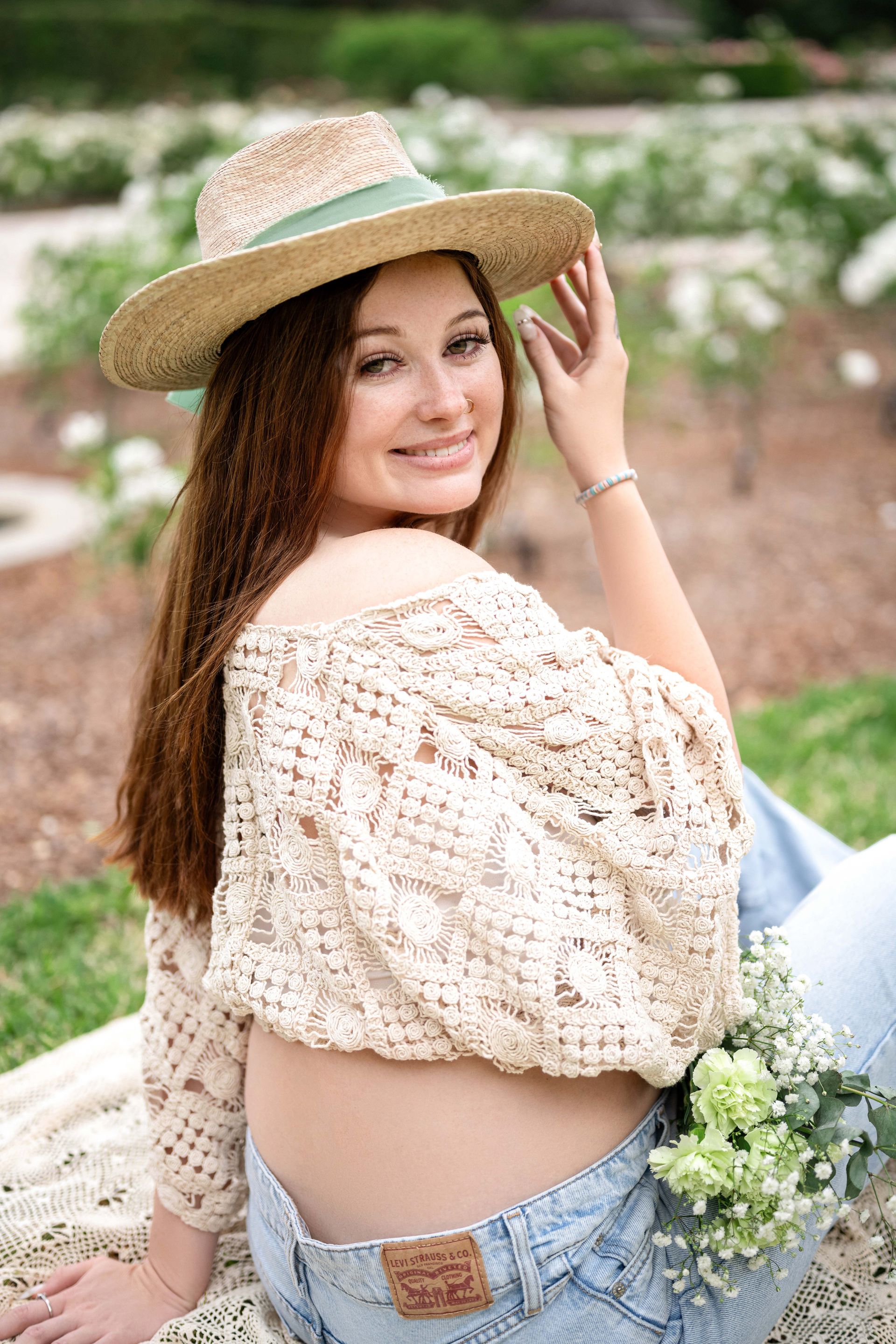 Woman wearing a straw hat, crocheted top, and jeans, smiles outdoors with flowers.