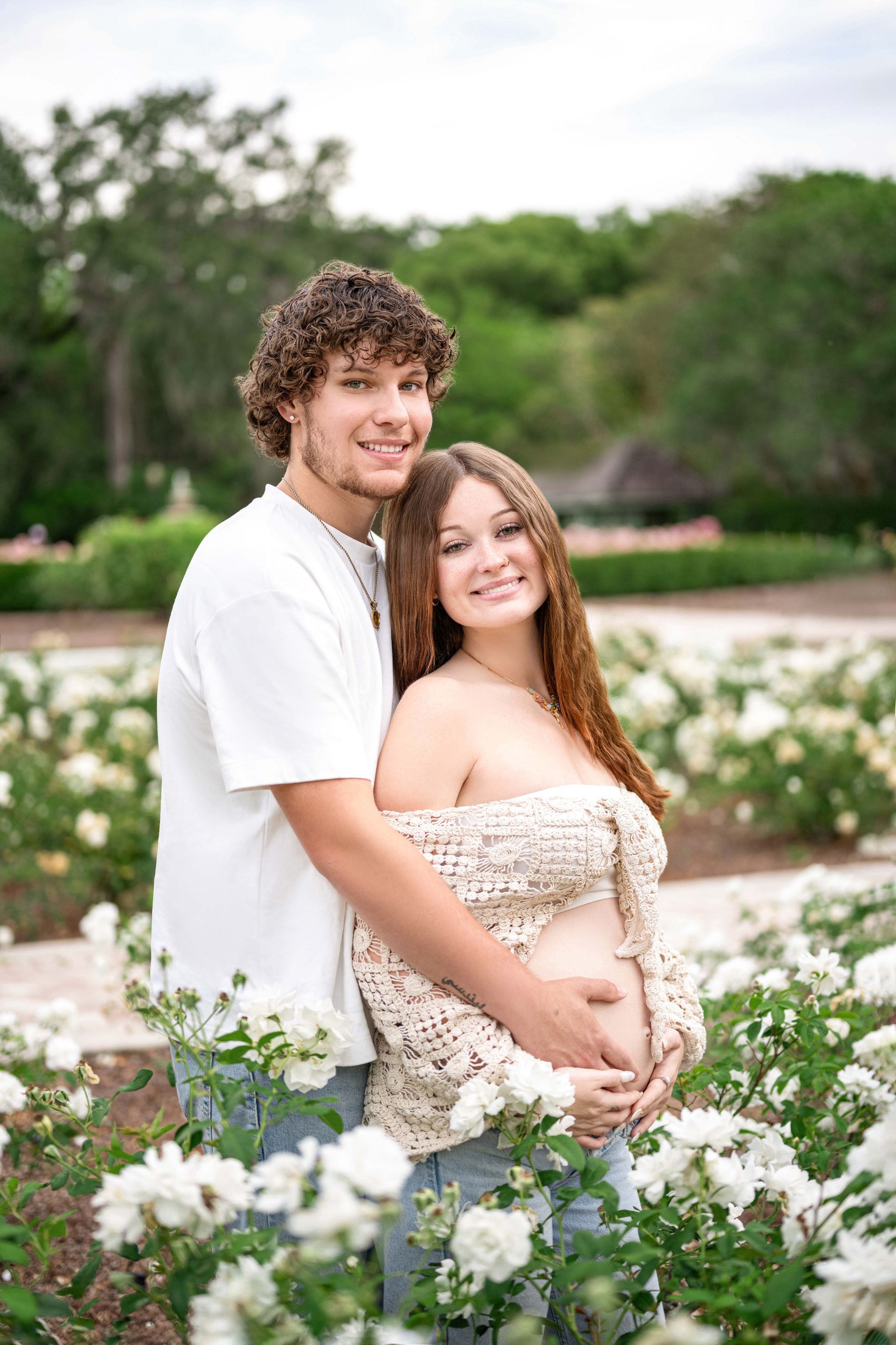 Couple in rose garden, man hugging pregnant woman, both smiling.