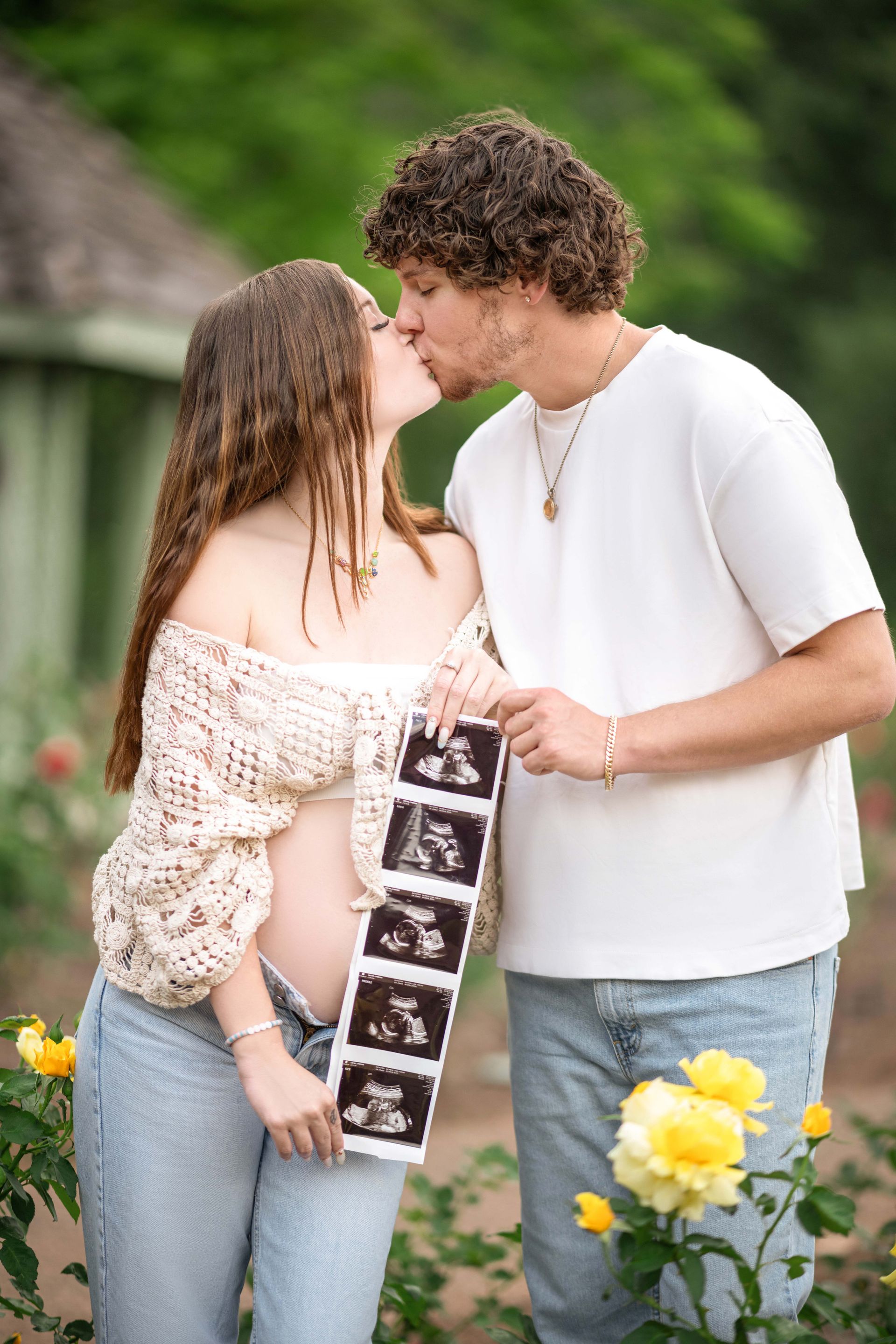 Couple kissing, holding sonogram images, with baby bump visible. Outdoor garden setting, yellow roses.