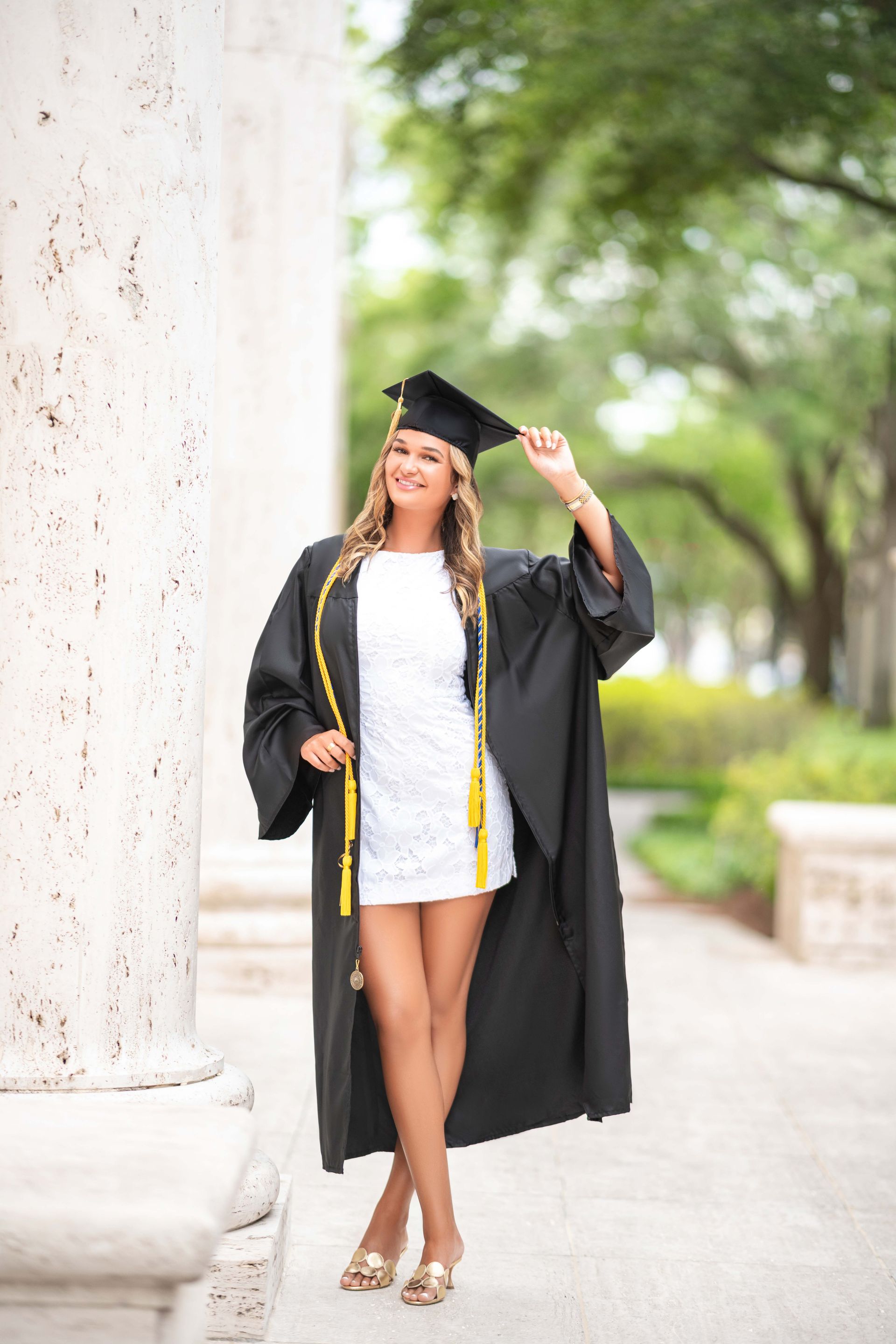 Woman in graduation gown and cap, smiling near a column, gold shoes, outdoor setting.