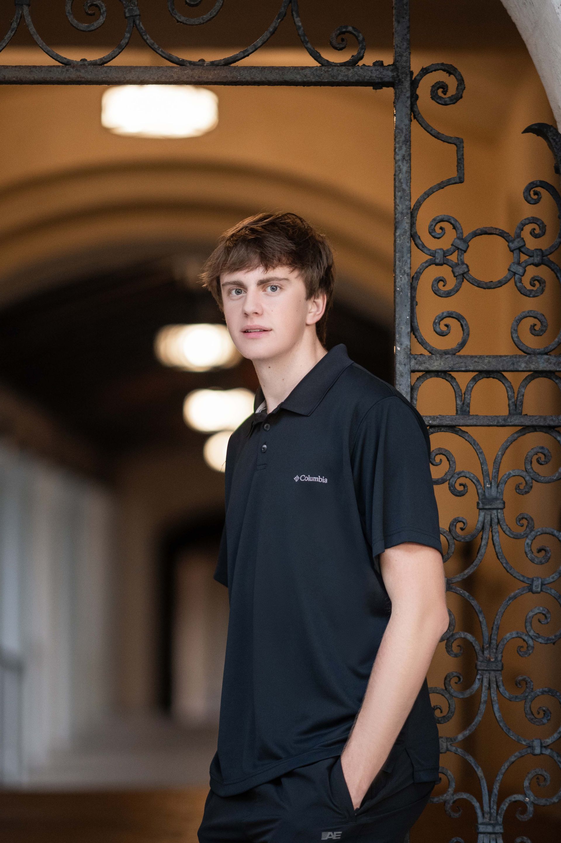 Young man in black polo shirt leans against an ornate gate in a long hallway.