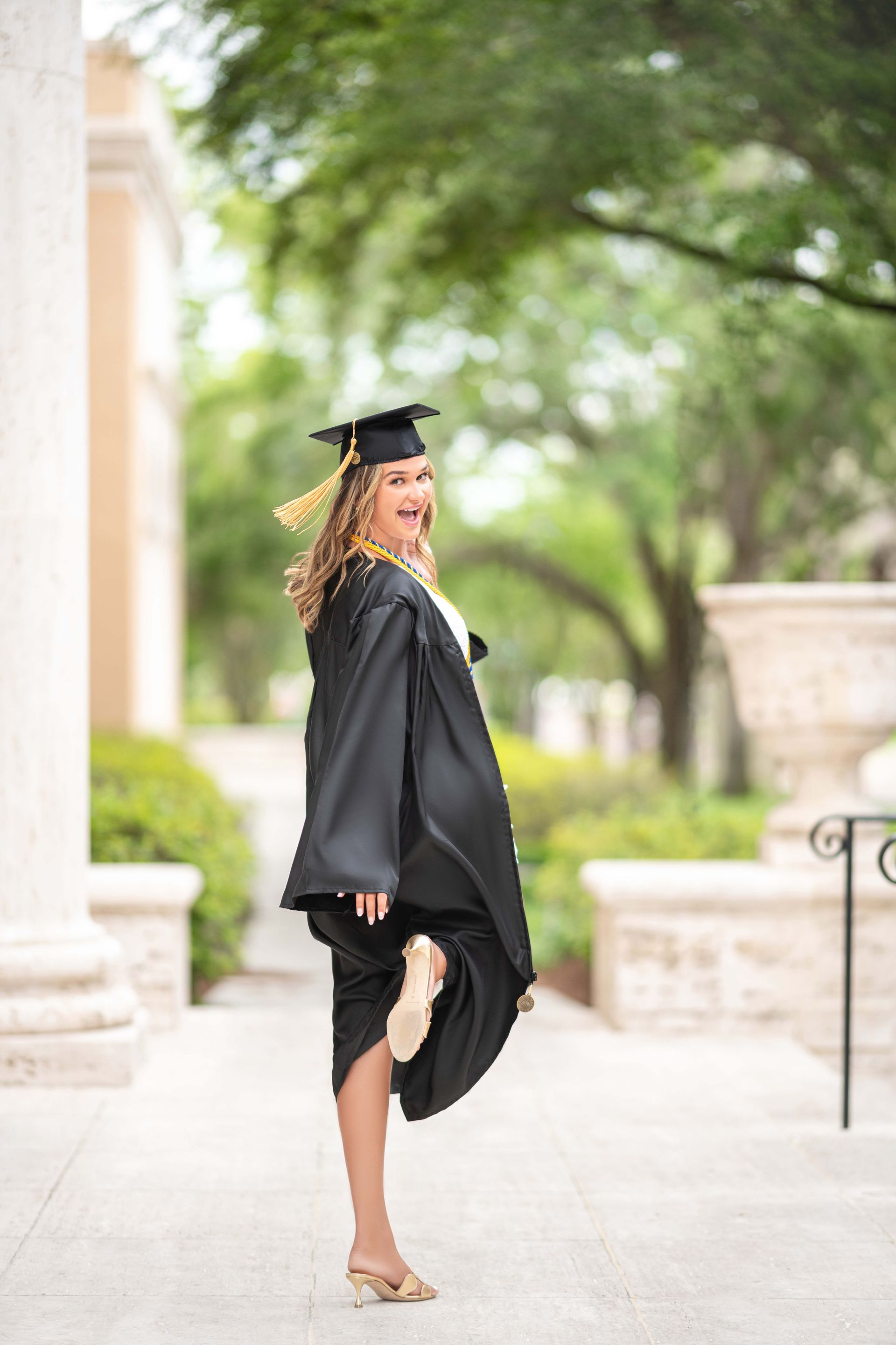 Woman in graduation gown, looking back and smiling, holding up one leg in a sunny outdoor setting.