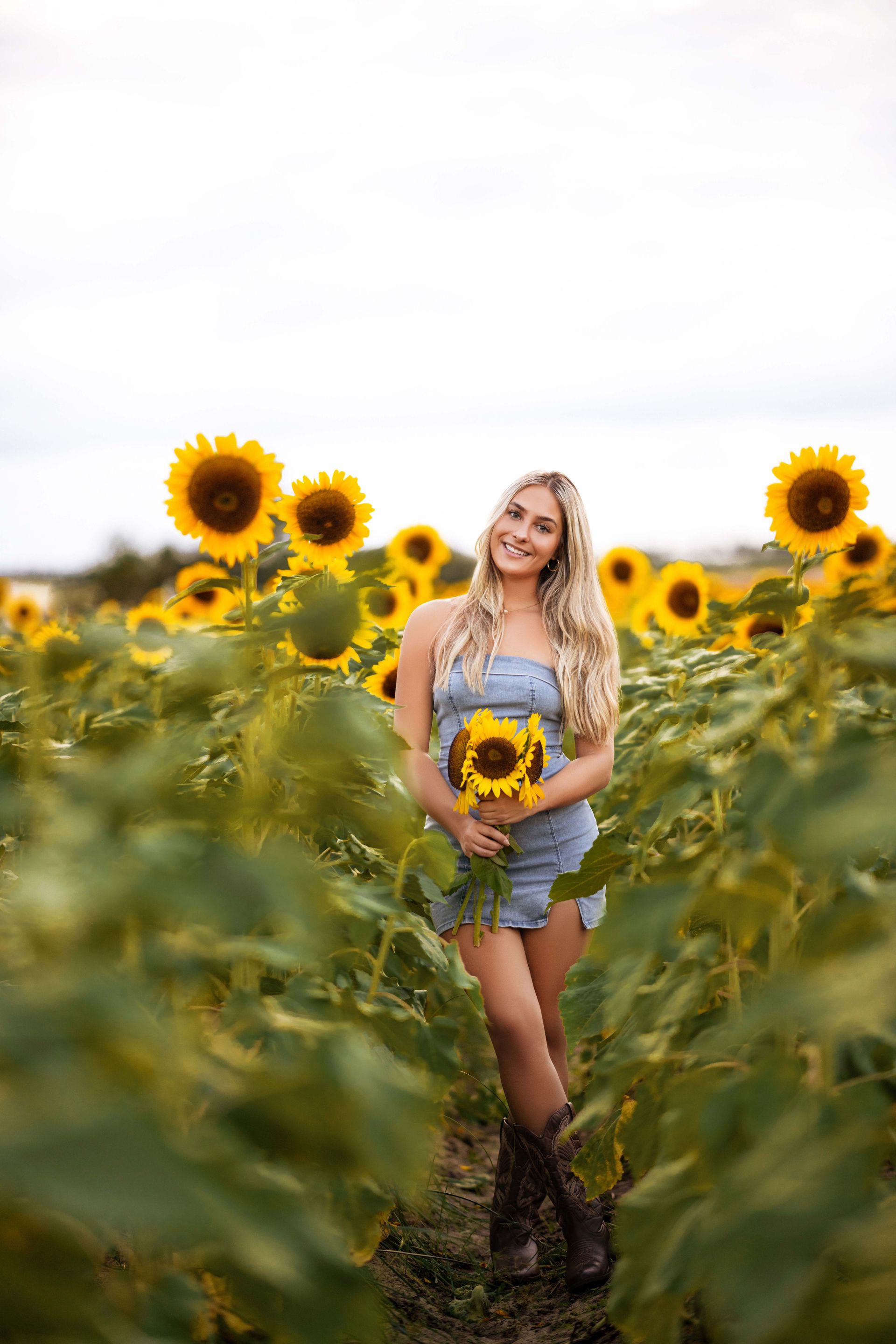 Blonde woman in denim overalls and boots smiles, holding sunflowers in a sunflower field.