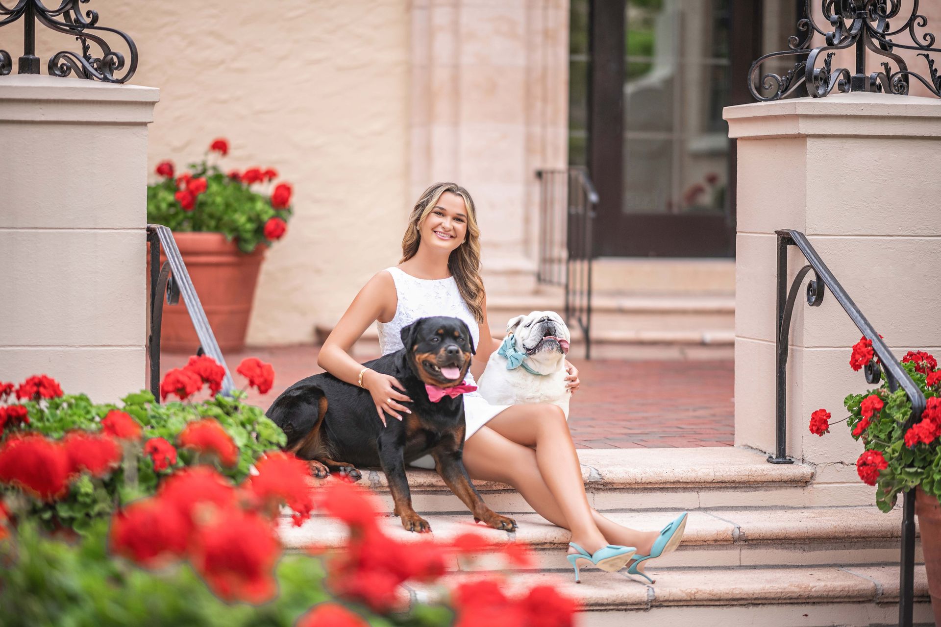 Woman sits with two dogs on steps with flowers. She smiles.