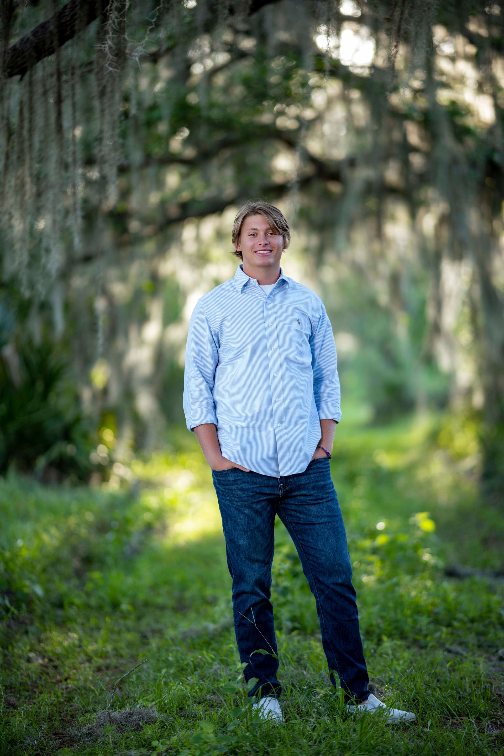 Young man stands in a grassy forest clearing, smiling, wearing a blue striped shirt and jeans.