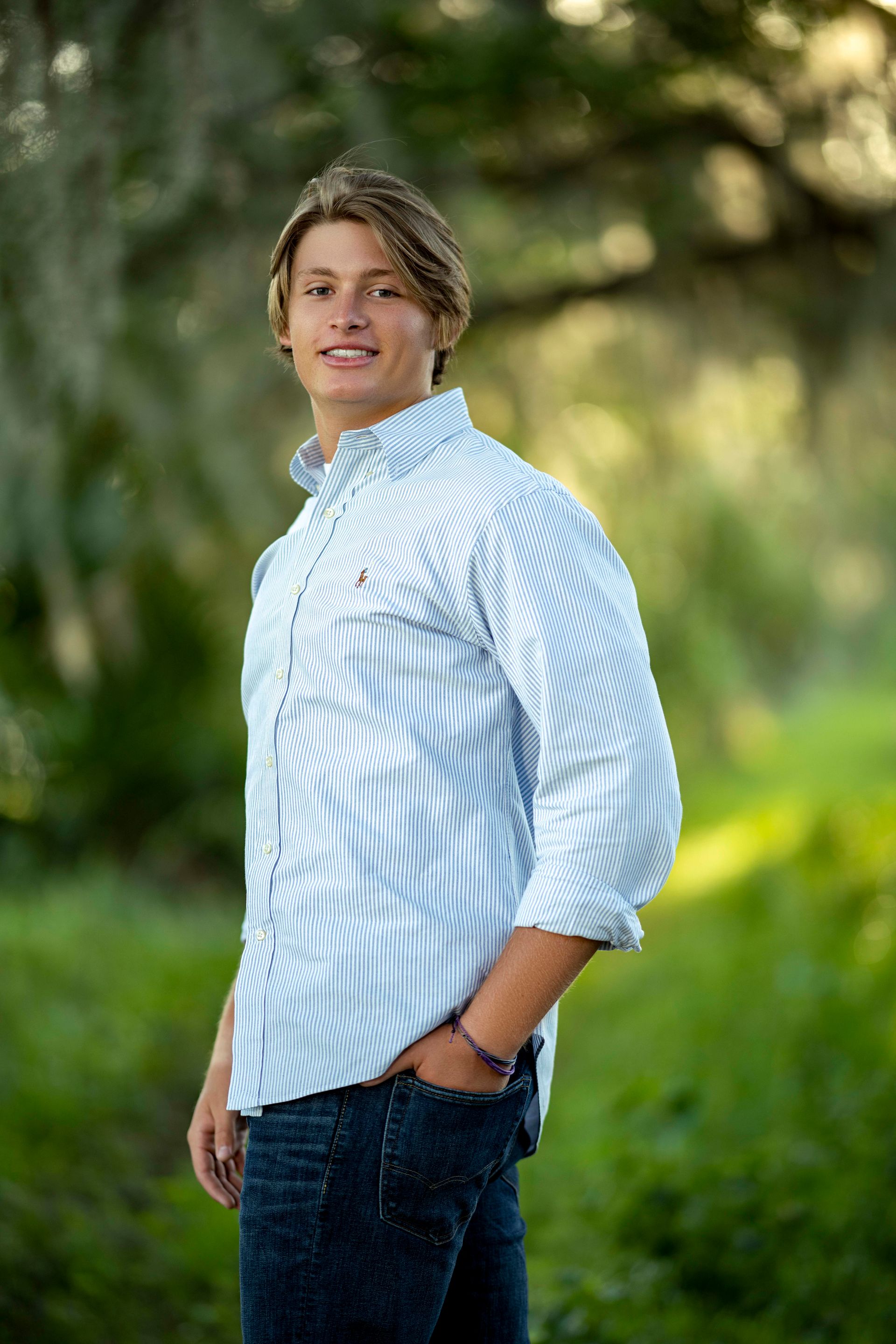 Blond young man in a blue and white striped shirt stands outside with a hand in his pocket smiling.