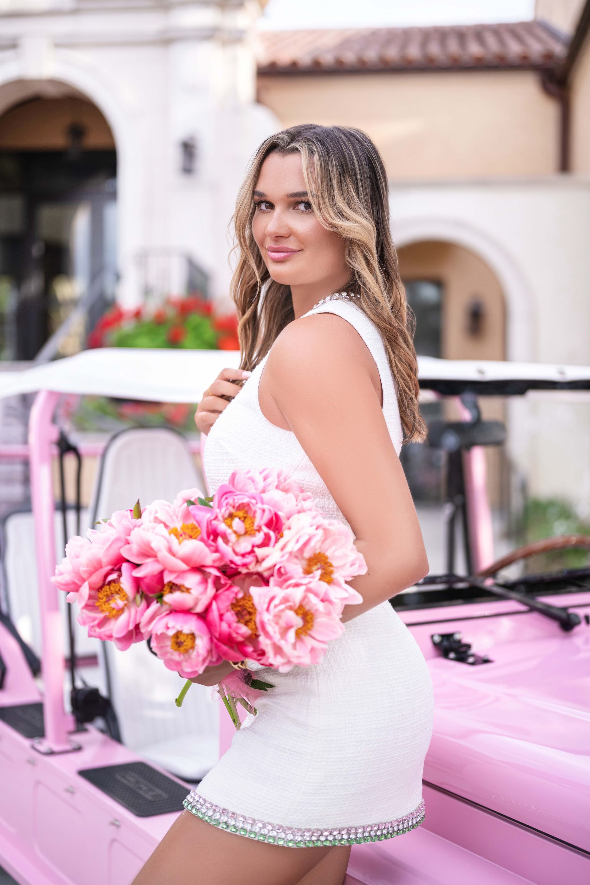 Woman in white dress holds pink flowers, posing by a pink golf cart.