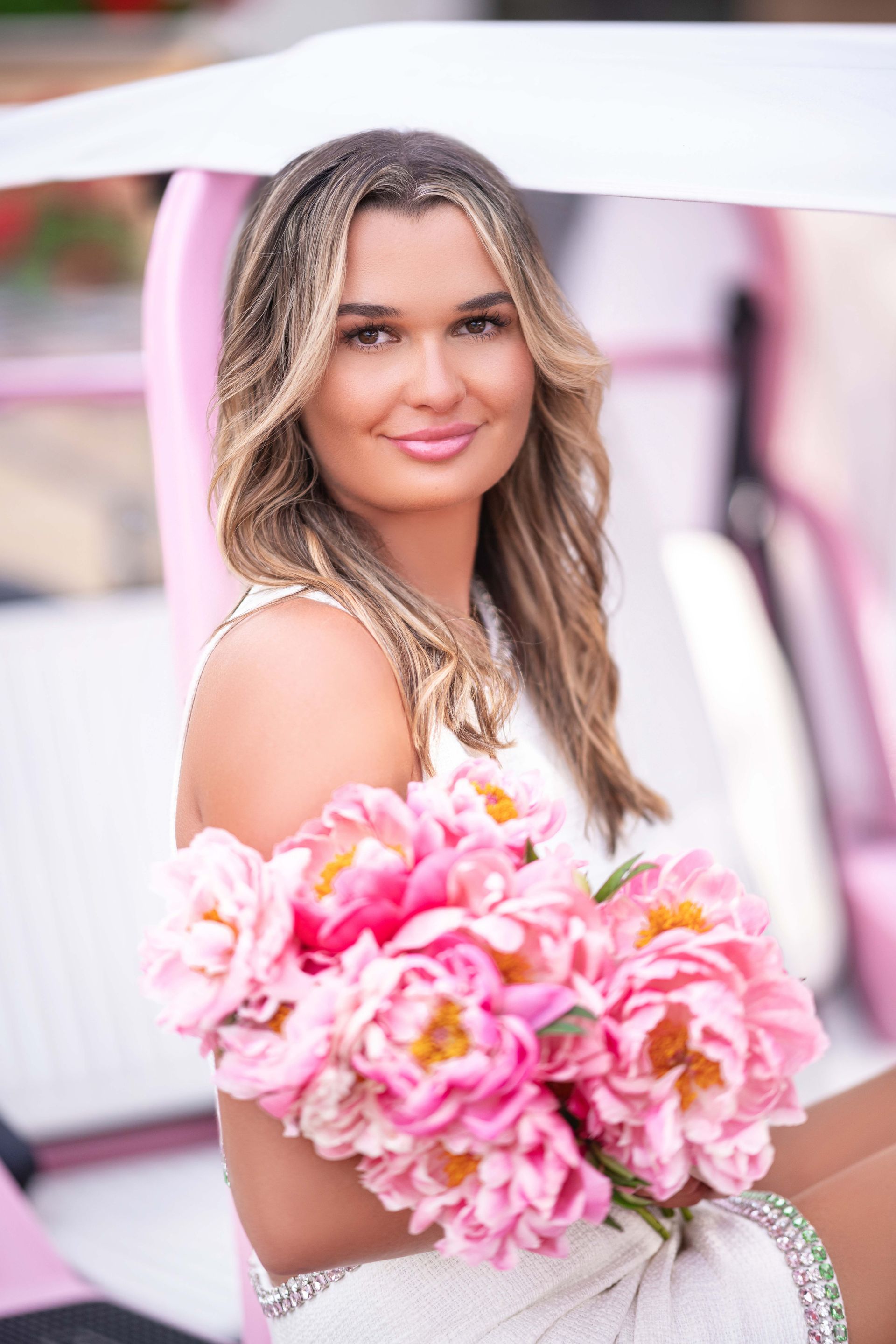 Woman in white dress holds pink flowers, seated in pink golf cart, smiling.