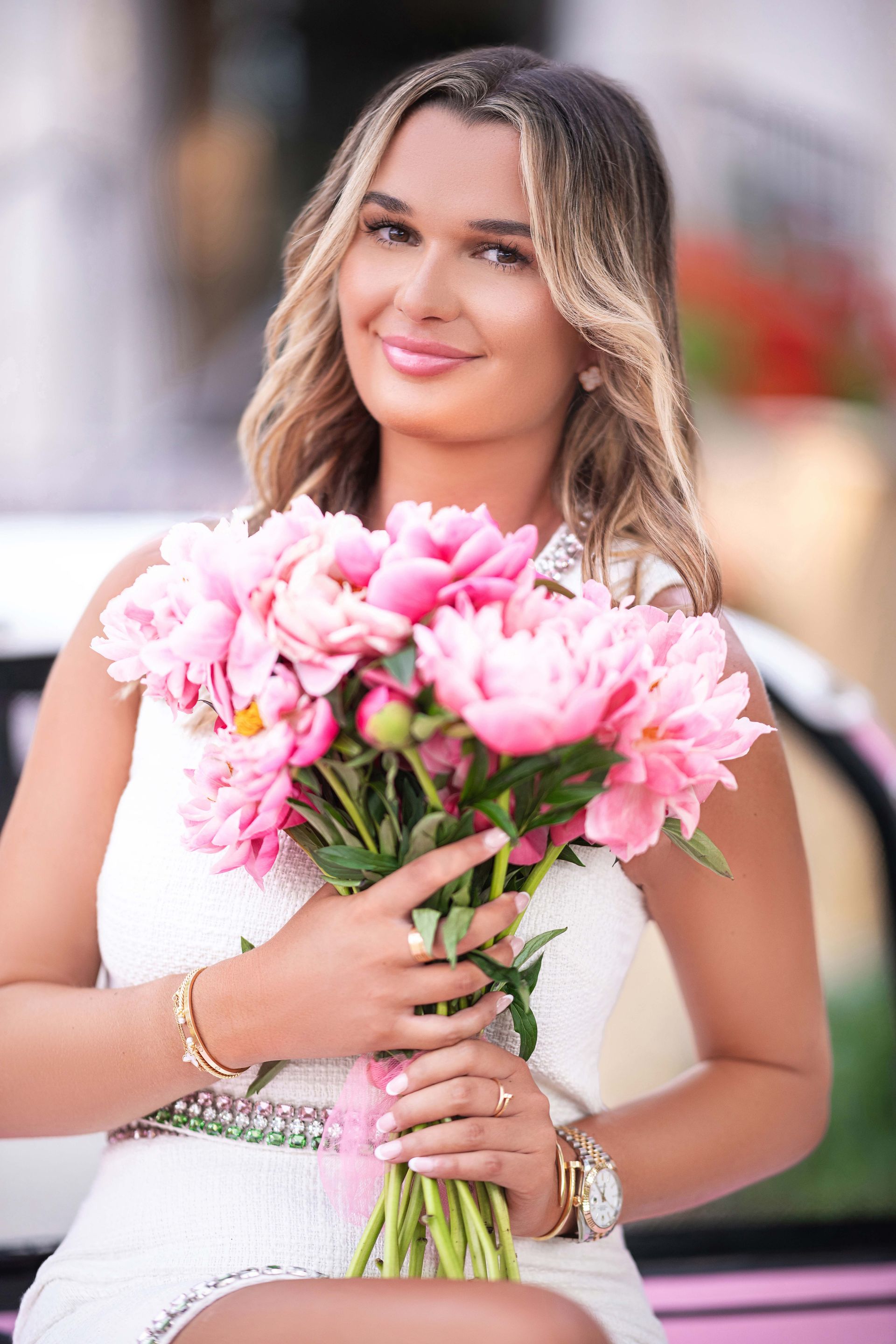 Woman in white dress smiles, holding pink peonies. Outdoors, near a pink golf cart.