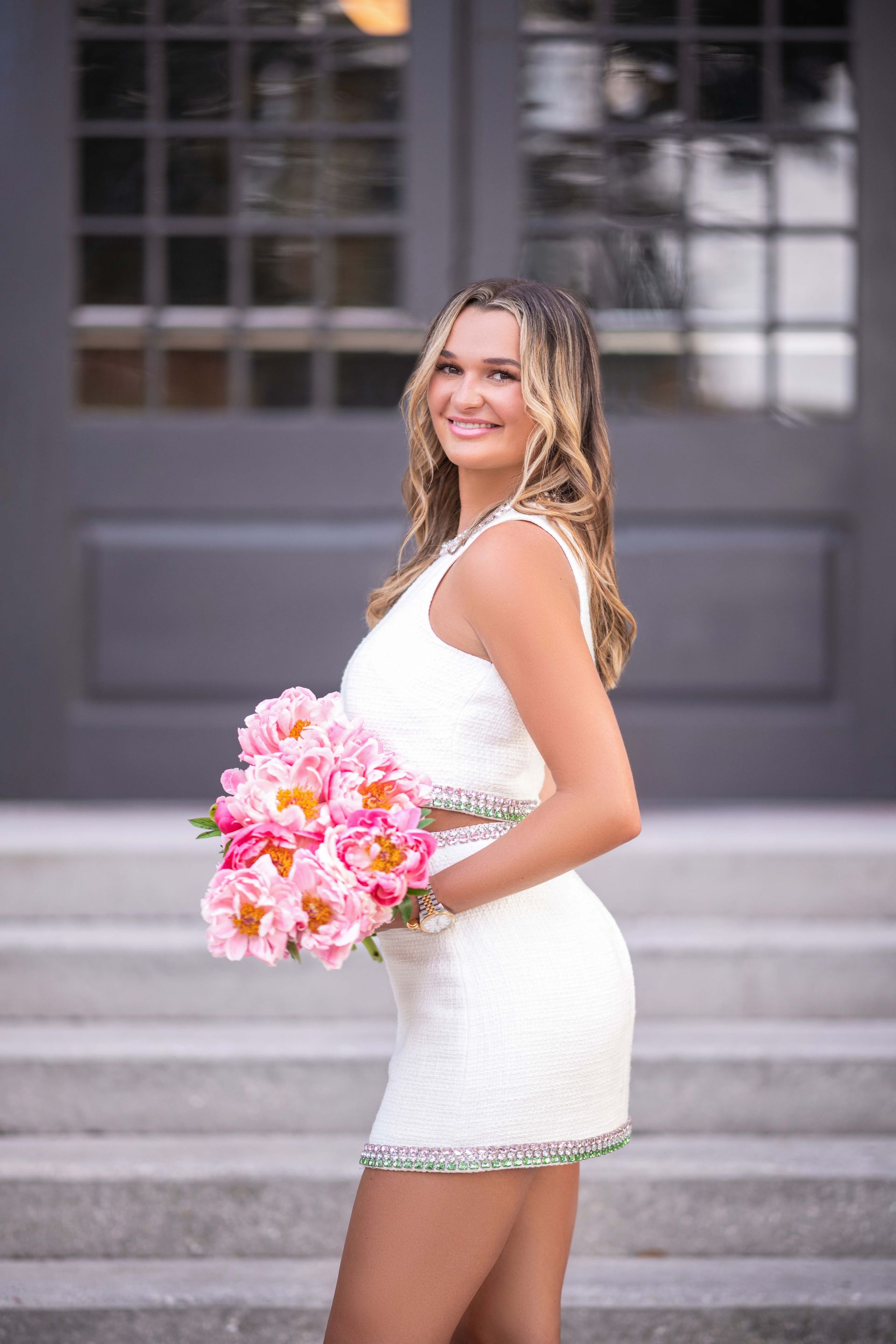 Blonde woman in white dress holding pink flowers, smiling outdoors in front of gray building.