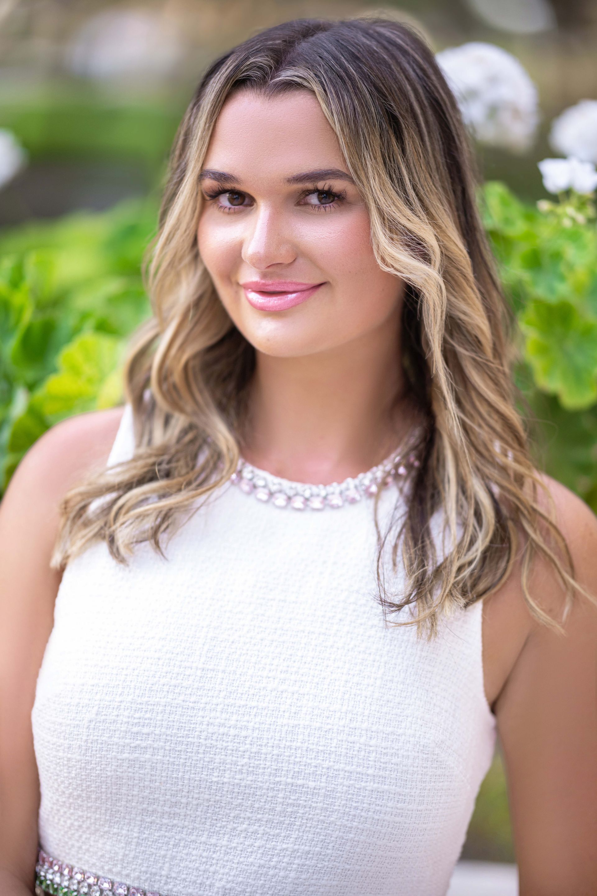 Woman with blonde-highlighted hair smiles, wearing a white dress, posing outdoors near green plants.