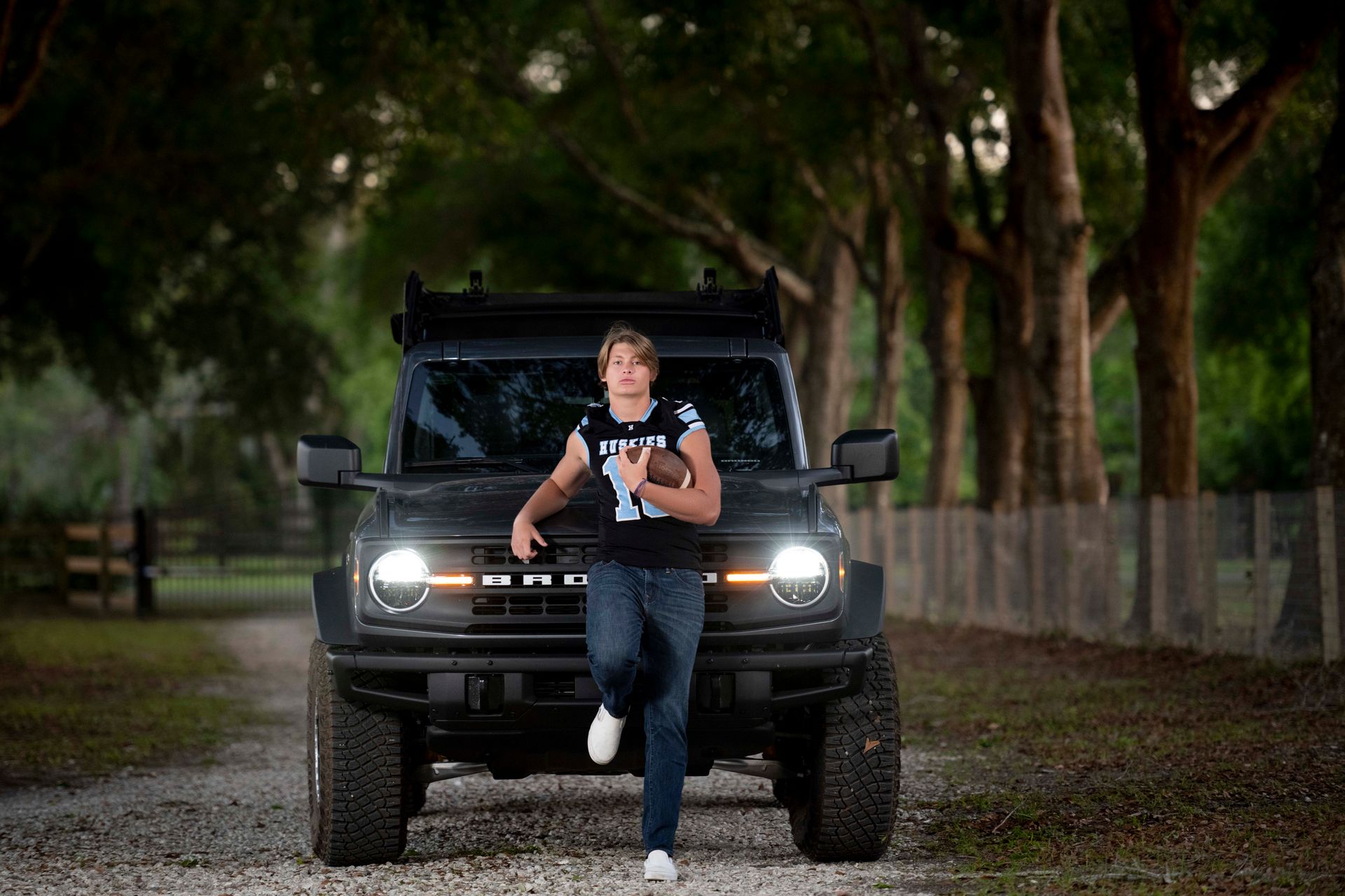 Teenage boy leans against a black Bronco, holding a football, on a dirt road.