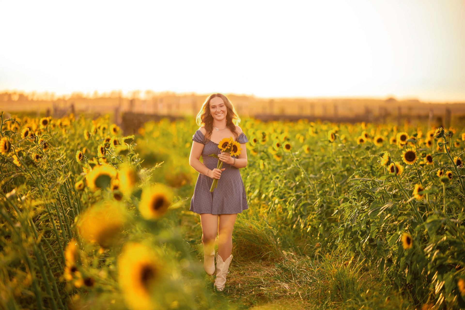 Woman in blue dress and cowboy boots walks through a sunflower field, holding sunflowers, smiling.