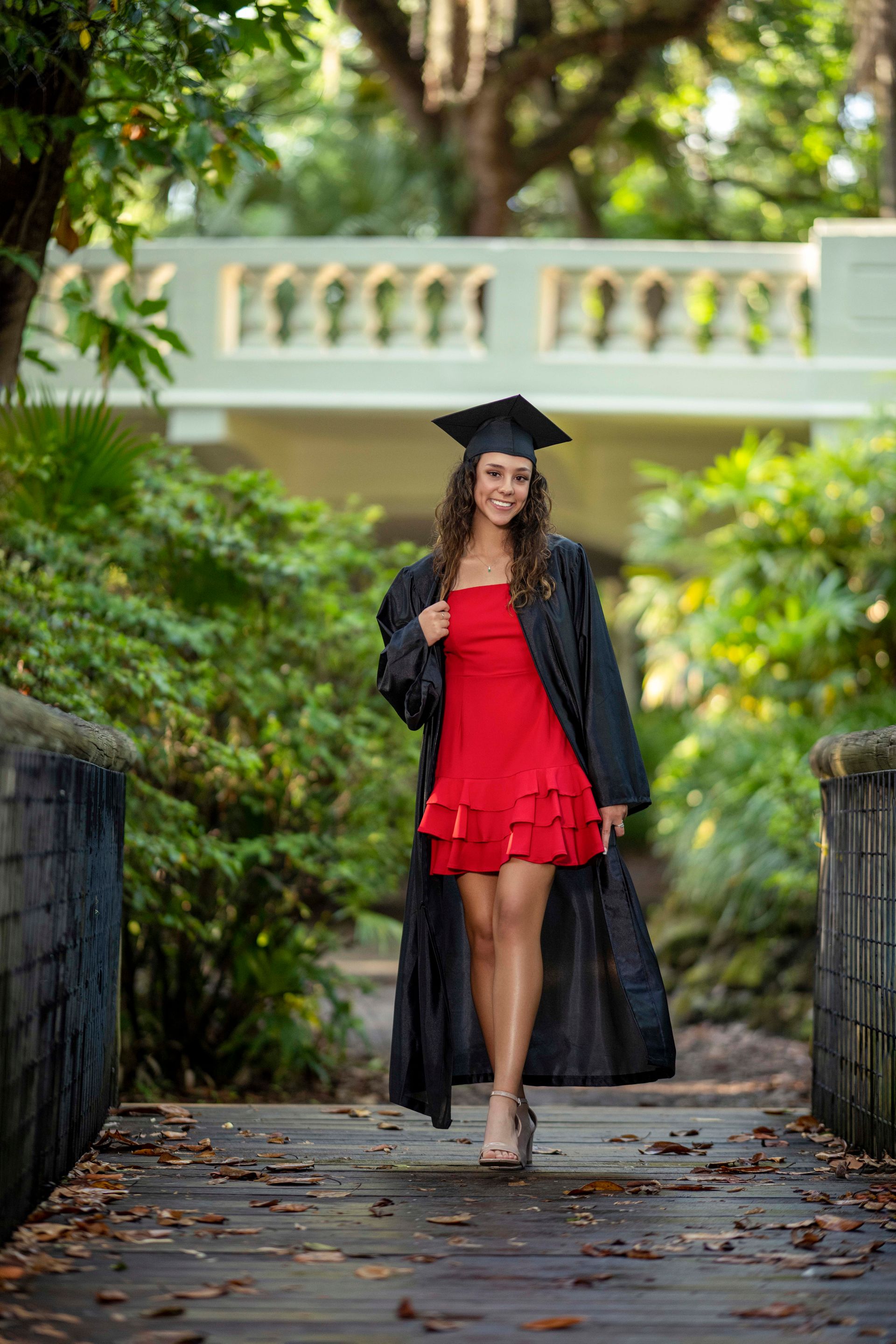 Woman in graduation cap and gown, red dress, walking on a bridge.