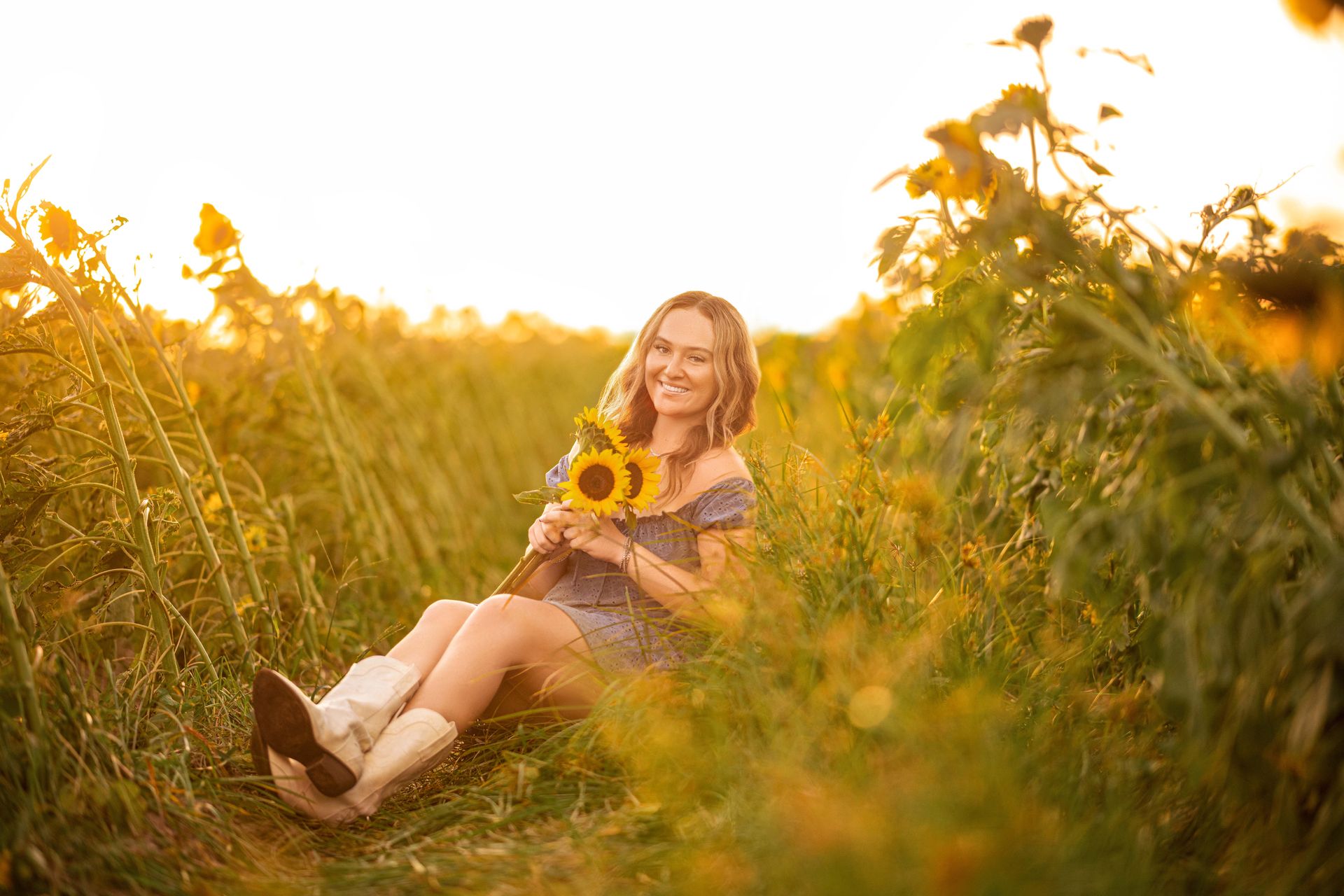 Woman sitting in a sunflower field at sunset, holding sunflowers and smiling. She wears a blue dress and cowboy boots.