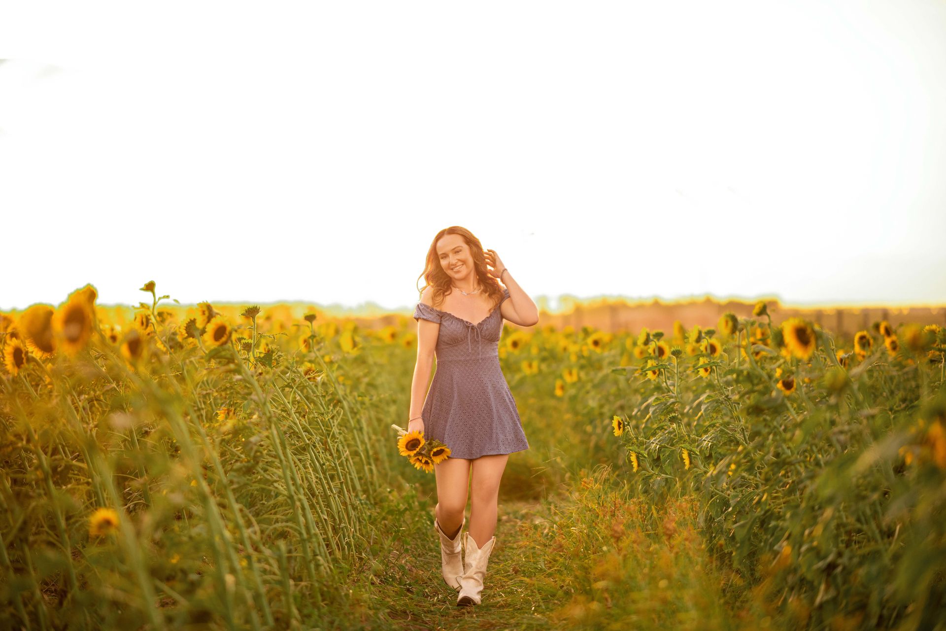 Woman in a blue dress smiles, walking through a sunflower field, holding flowers, with a sunset in the background.