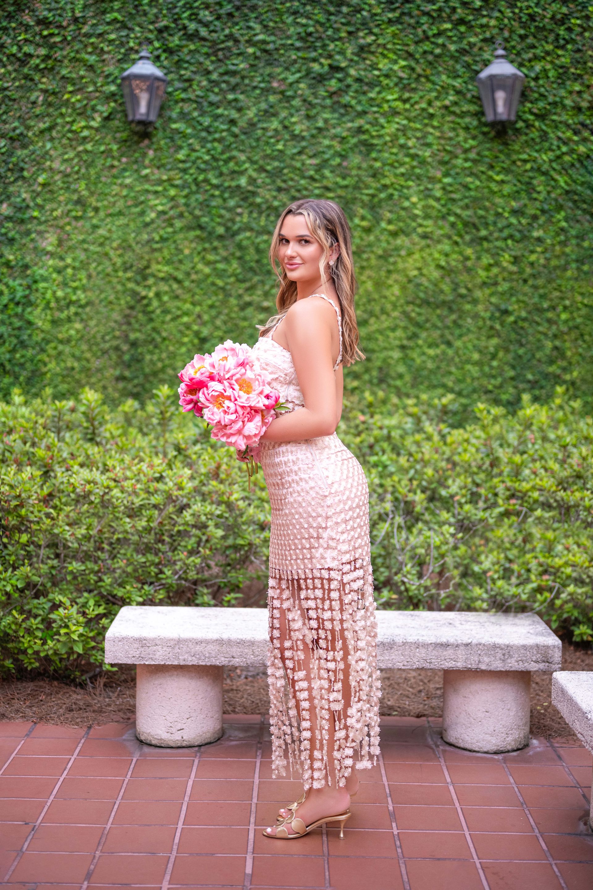 Woman in a pink gown holding flowers, standing near a bench and a green wall.