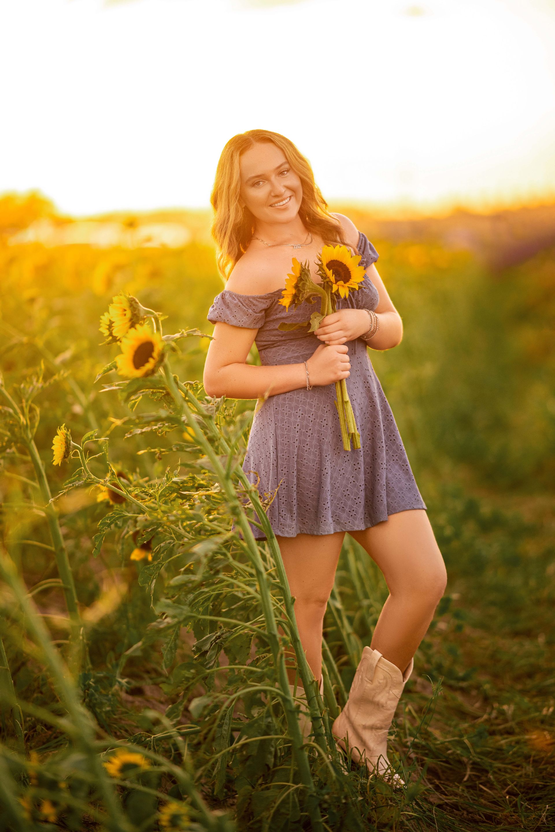Woman in a blue dress holding sunflowers, standing in a sunflower field at sunset.