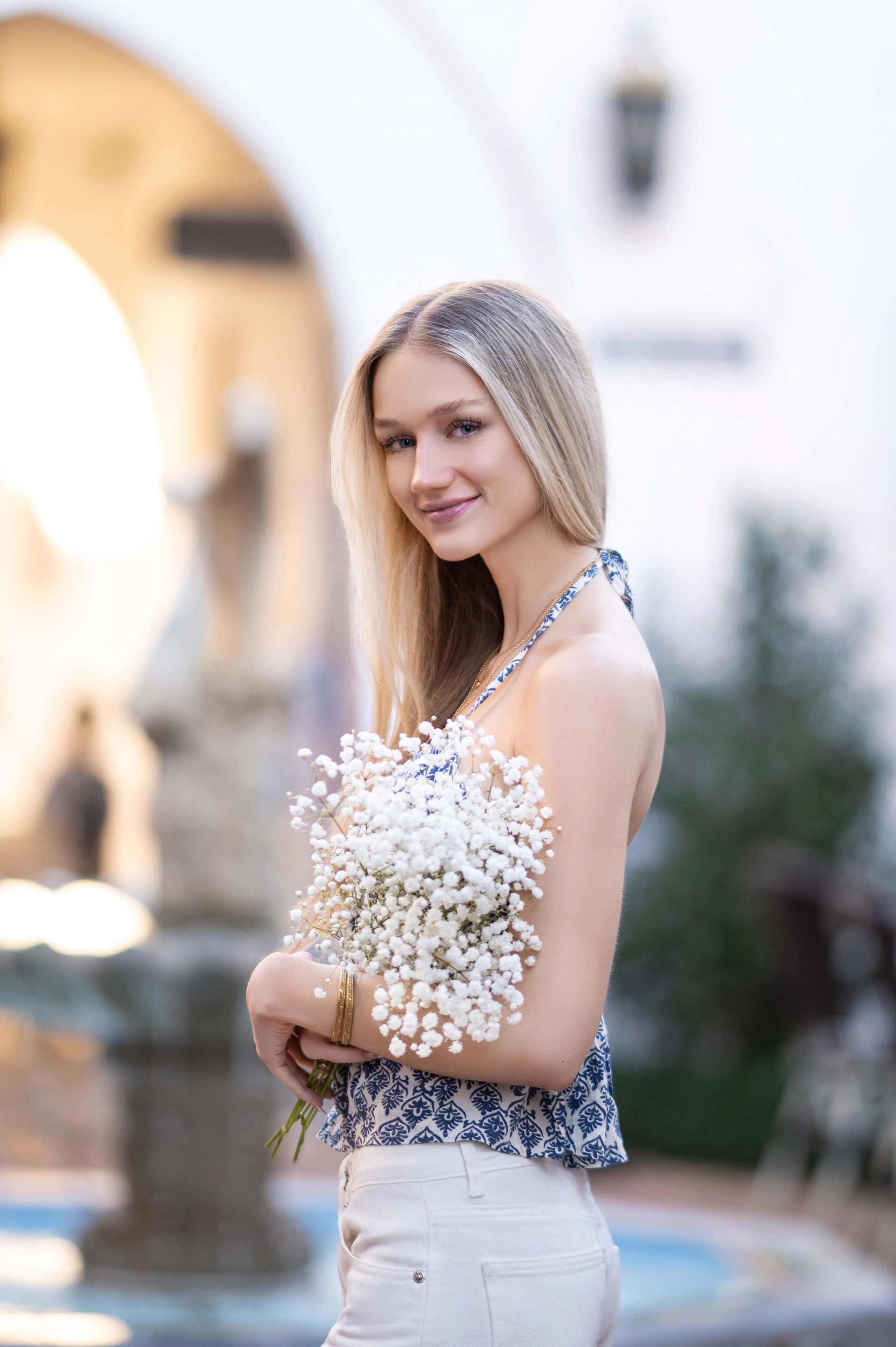 Blonde woman smiling, holding white flowers, wearing patterned top, standing outdoors near fountain.